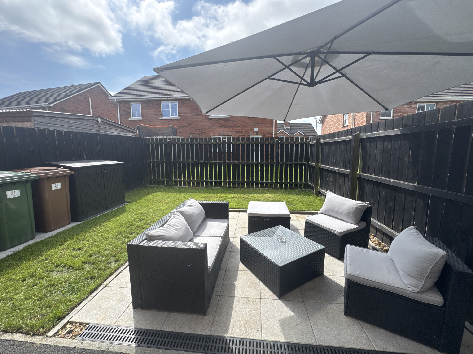 This is a forensic, matter-of-fact description of an outdoor patio scene in Craigavon, United Kingdom. The image depicts a small, fenced-in backyard area. In the foreground, a black rattan furniture set is arranged on a tiled patio. This set includes a sofa with light grey cushions, a coffee table with a glass top, a square ottoman with a light grey cushion, and an armchair with light grey cushions. The furniture is positioned towards the right side of the frame. Behind the furniture, a dark wooden fence encloses the yard. Beyond the fence, brick houses with grey tiled roofs are visible, indicating a residential area. To the left of the patio, a strip of green lawn runs along the fence line. Three wheelie bins, one green and two brown, are placed along the edge of the lawn, near a dark brown storage shed. The sky above is blue with scattered white clouds, suggesting daytime. A large, light grey cantilever umbrella is partially visible, extending over the patio area, providing shade. The overall scene is static and appears to be a typical domestic garden setting. There are no people or animals present in the image.