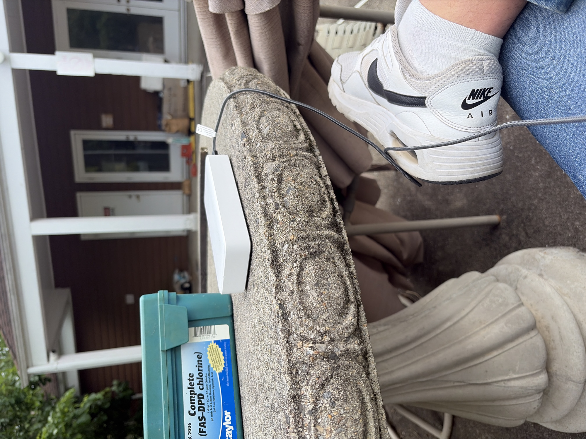 The image shows a close-up view, likely taken outdoors, with a combination of natural and man-made elements. A weathered, ornate concrete table is in the foreground, upon which a white electronic device and a blue container labeled "Taylor Complete (FAS-DPD chlorine)" are placed. A black cable is connected to the electronic device. A person wearing light blue jeans and white Nike Air sneakers with black swooshes is visible. The background shows a two-story house with white pillars and a brown exterior. There are green trees. The location is likely in Edison, United States of America.
