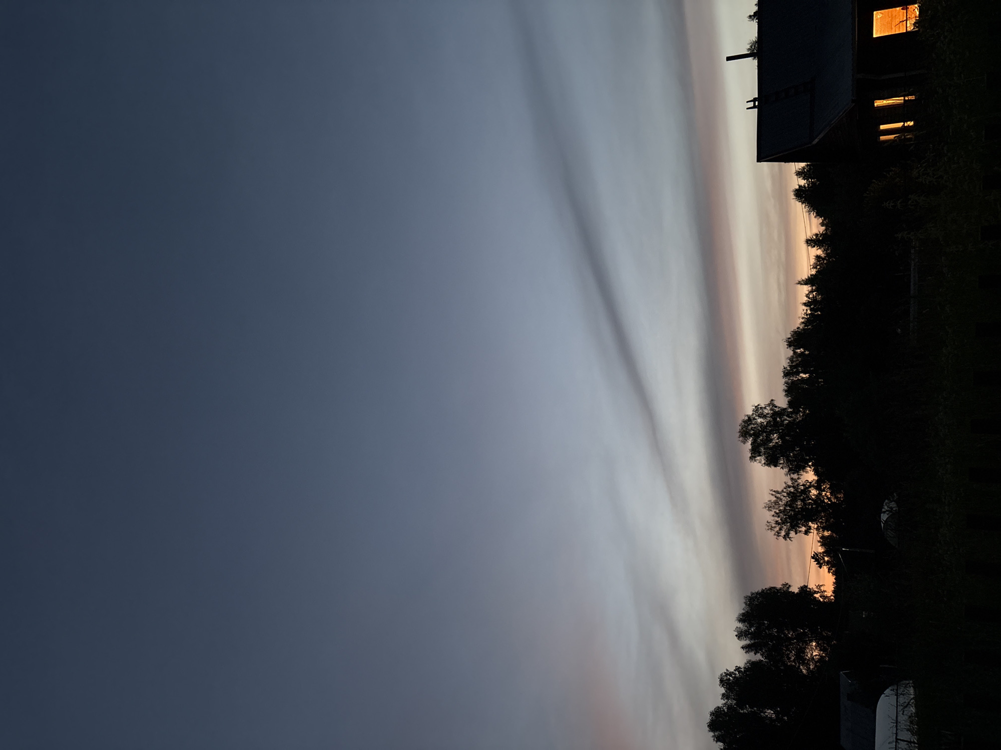 This is a vertical, nighttime photograph taken outdoors in Dubrovino, Russia. The scene captures a twilight sky with streaks of light, likely from clouds catching the last rays of the sun. On the right side of the image, a dark silhouette of trees borders the sky. A dark-colored building with illuminated windows is visible on the far right, suggesting indoor activity. The overall mood is serene and peaceful, indicative of the end of the day. The lighting suggests it is either dawn or dusk. No people are visible in the image, and there is no visible text.