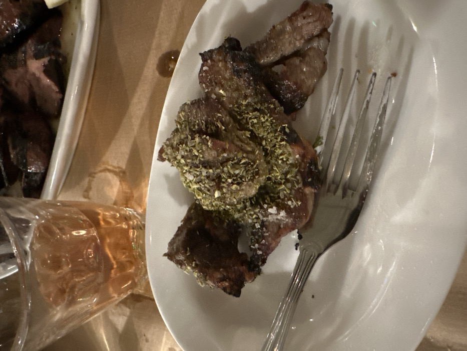 The image captures a close-up, overhead view of a dining scene, likely indoors in Nea Erithrea, Greece. The primary subject is a serving of cooked meat, possibly grilled lamb chops or steak, presented on a white oval plate. The meat pieces are irregularly shaped, with visible charring and a reddish-brown interior. They are generously seasoned with a dense layer of dried green herbs, resembling oregano, and scattered white crystals, likely sea salt. A silver four-tined fork rests on the right side of the plate, partially obscuring some of the meat.
To the left, a portion of another white rectangular dish containing more cooked meat, similar in appearance, is visible. Below this dish, a clear glass filled with an amber-colored liquid, possibly a drink like wine or a spirit, is present. The liquid shows reflections of light, and the glass itself appears to be slightly toppled or angled, with liquid partially visible. The entire arrangement rests on a brown or beige surface, which could be a table or placemat. There's a small, dark liquid spill or drop on this surface between the two dishes. The lighting is warm and somewhat dim, suggesting an evening meal or an indoor setting with ambient lighting. No people or visible text are present in the image.