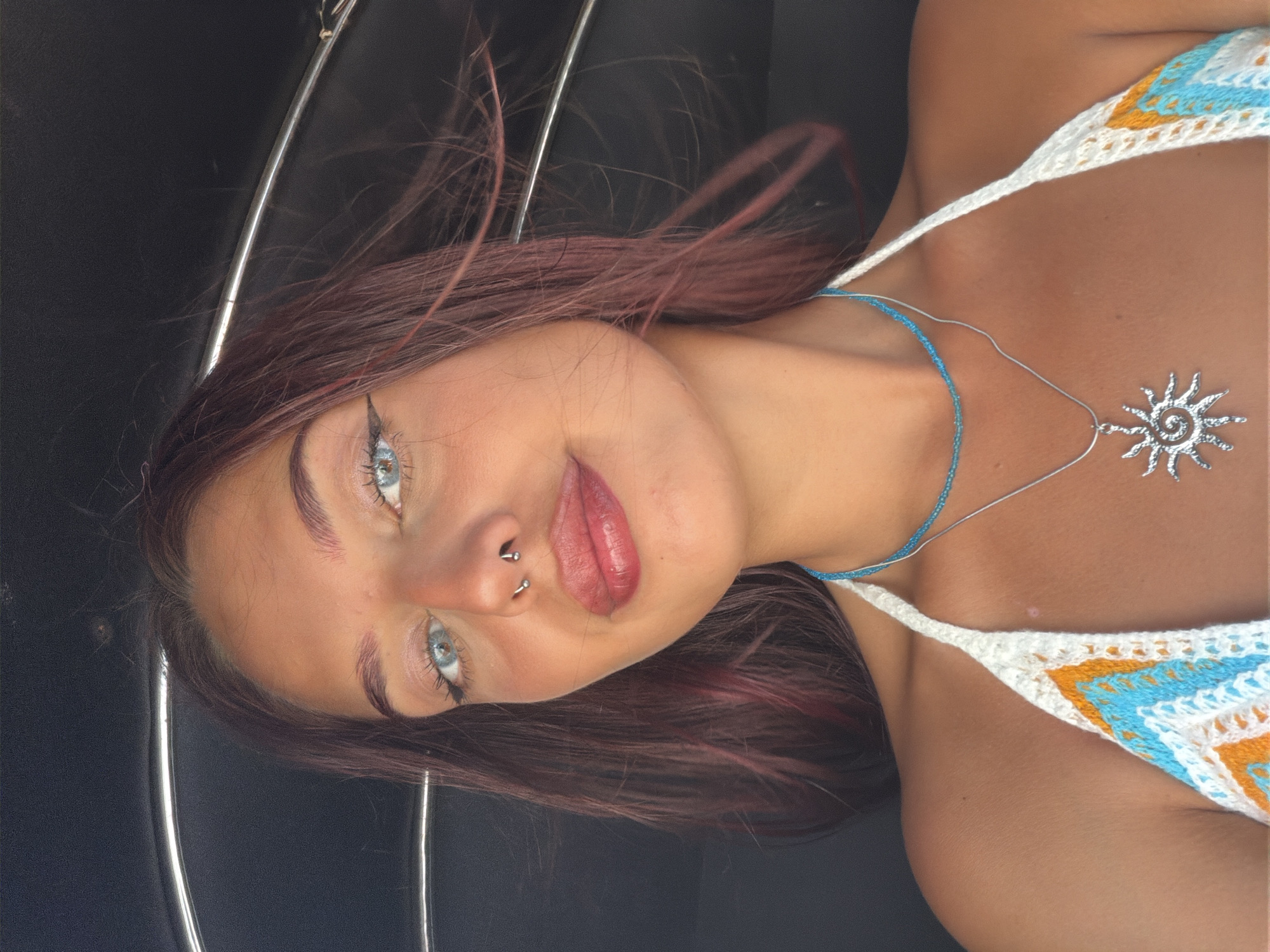 A young woman with light blue eyes, dark eyebrows, and auburn hair adorned with a septum piercing and a necklace, is shown in a close-up, frontal shot. She is wearing a white, crochet halter top with a blue and orange zigzag pattern. The background is a dark, blurry interior with a curved silver railing visible. The woman's hair is slightly windswept, suggesting an outdoor or moving environment. The image appears to be taken in Pottuvil, Sri Lanka, a location known for its beaches and surfing spots.