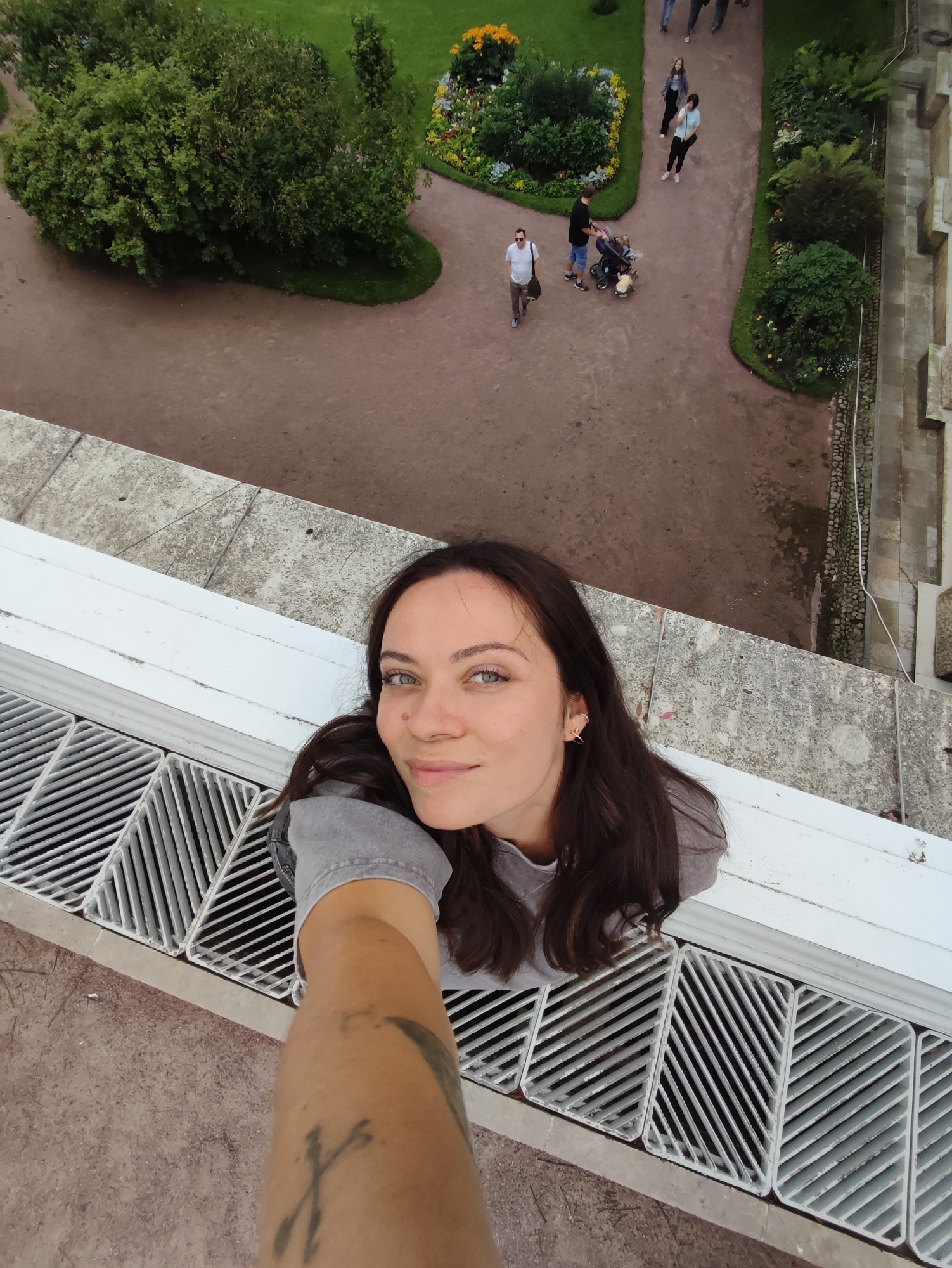 An overhead, selfie-style shot captures a young woman with long, dark brown hair and blue eyes, looking up at the camera from a lower vantage point. She is wearing a grey t-shirt. Her arm, with a visible tattoo, extends towards the camera. She is positioned on a white ledge, above a series of grey metal grates. Below her, a paved pathway winds through a park with lush green trees and flower beds. Several people are visible on the pathway: a man in a white t-shirt walks away from the camera, while another man pushes a stroller with at least one child. A couple walks further down the path, and another individual is partially visible. The environment appears to be an outdoor public space, consistent with a park in the city of Pushkin, Russia, possibly within the grounds of Catherine Palace, known for its gardens and pathways. The lighting suggests an overcast day.