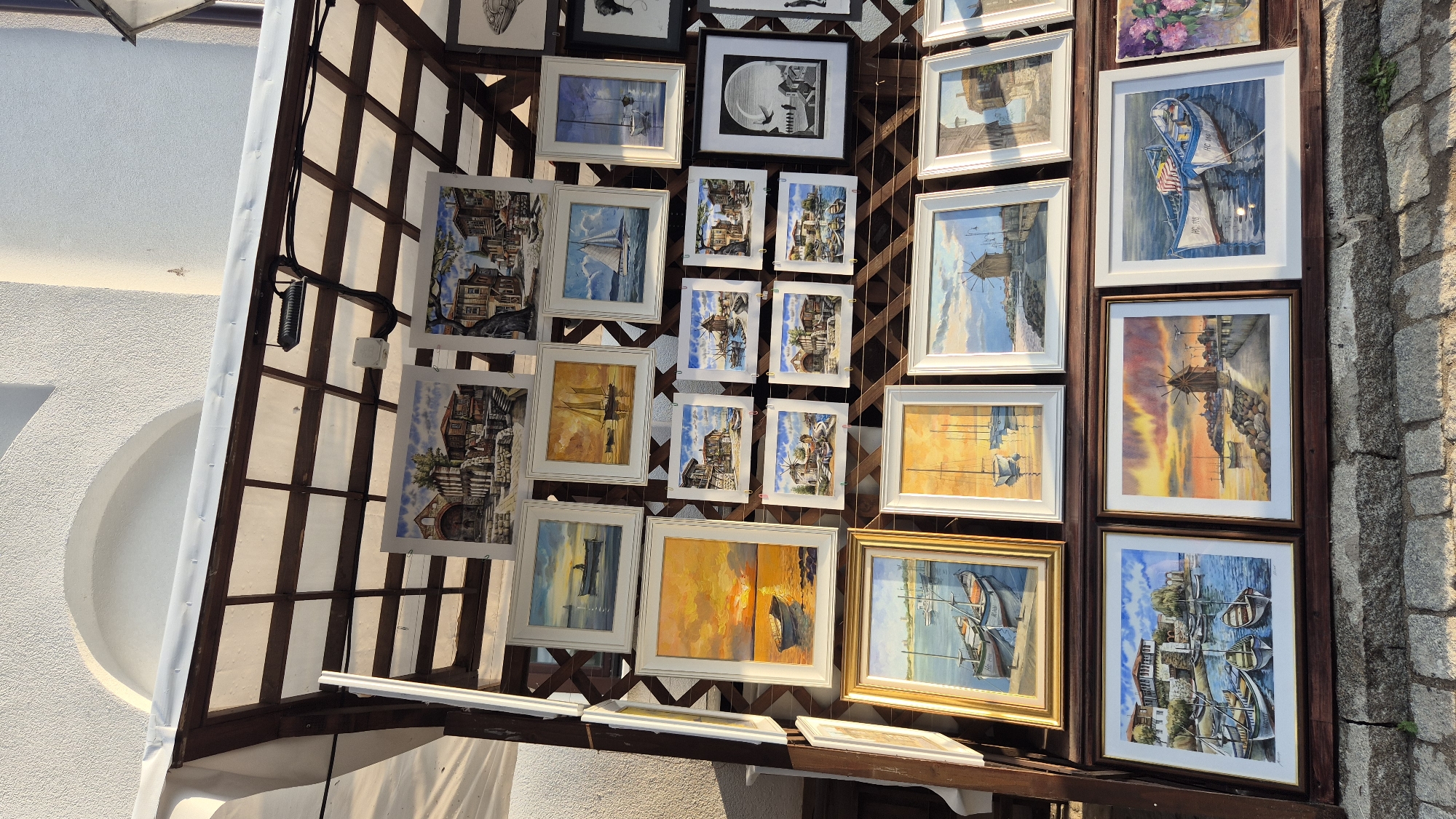 The image depicts an outdoor art display in Nesebar, Bulgaria, showcasing numerous framed paintings. The art is mounted on a wooden lattice structure, likely part of a market stall or gallery setup, with a white textured wall and a white awning overhead. The paintings primarily feature maritime scenes and architectural views of Nesebar's old town, characterized by traditional wooden houses and cobblestone streets, as well as boats, harbors, and seascapes. The artwork varies in size and framing, with some being watercolors and others appearing to be oil paintings. The lighting suggests it is either early morning or late afternoon, with warm sunlight illuminating the scene. There are no people visible in the frame.