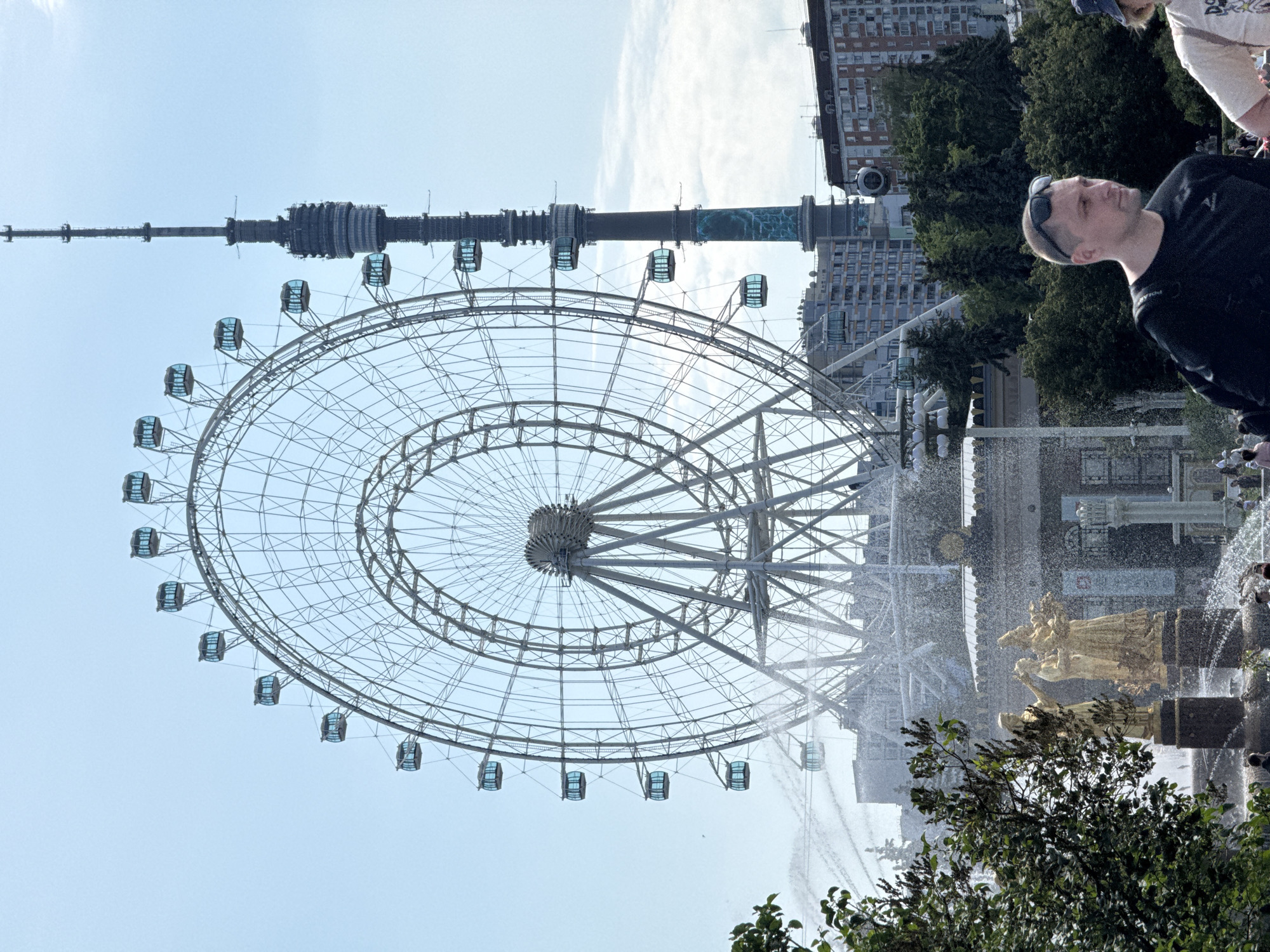 This is a medium shot, taken outdoors on a sunny day, featuring the Moscow Ferris Wheel and Ostankino Tower in Moscow, Russia. In the foreground, a man wearing sunglasses and a black t-shirt is looking to his right. To his right, another person, mostly out of frame, is wearing a light-colored t-shirt and a blue hat. Behind them, a large, light-colored Ferris wheel dominates the background, with several of its blue-tinted cabins visible. To the right of the Ferris wheel, the Ostankino Tower, a tall, dark structure, rises into the clear blue sky. In the middle ground, a fountain sprays water upwards, with golden statues of people visible in the center of the fountain. In the far background, several buildings are visible, including apartment buildings and a large, low-rise building with columns. Green trees are scattered throughout the scene, particularly in the foreground and middle ground.