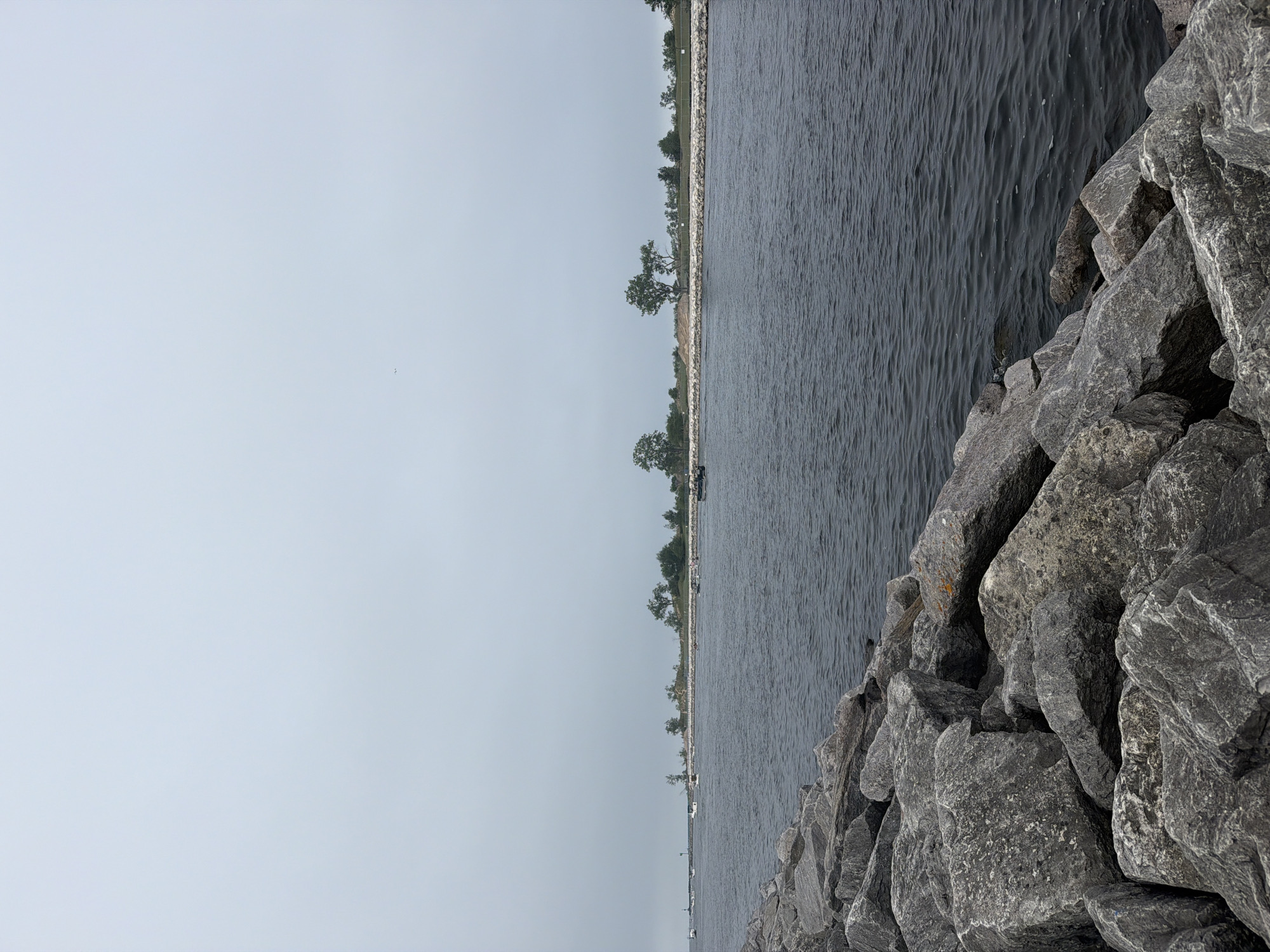 The image shows a wide body of dark water with a rocky breakwater in the foreground. The water extends towards a distant, tree-lined shore with several boats visible on the water. The sky is overcast and pale gray. The scene is likely a harbor or channel entrance, and given the prompt, is situated in Muskegon, Michigan, United States of America. There are no people clearly visible in the image. The overall impression is one of a calm, overcast day at a waterfront location.