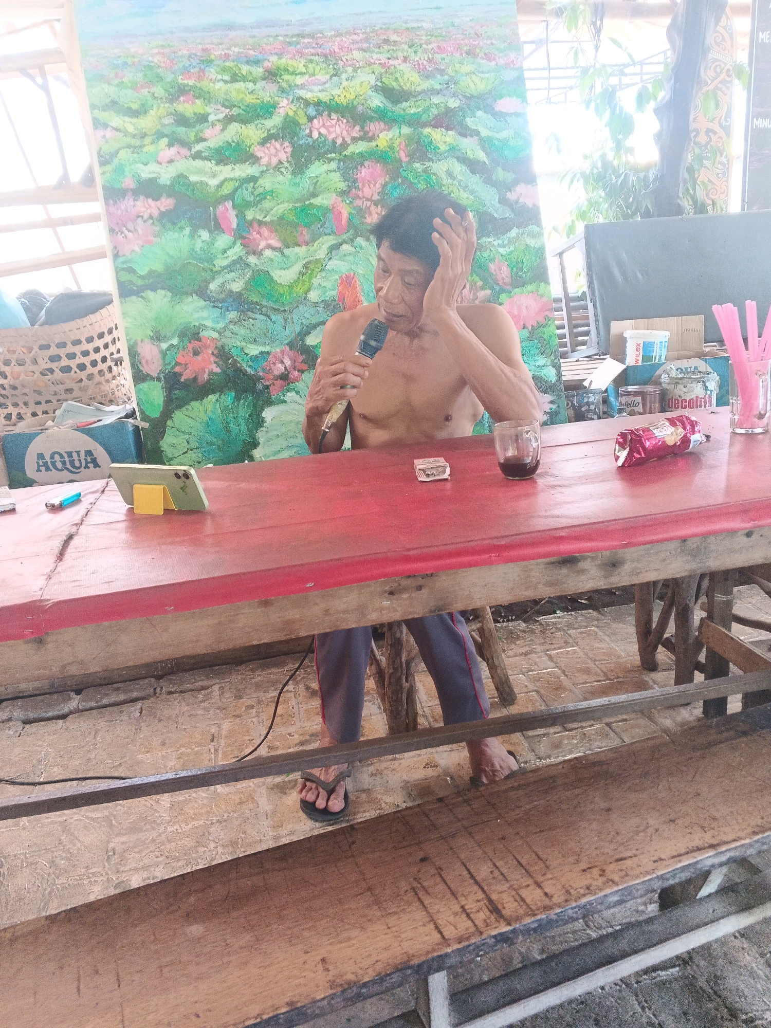 A shirtless man, with his hand on his head, sits at a red wooden table. He holds a microphone and appears to be speaking. In front of him on the table, there is a smartphone on a yellow stand, a lighter, a glass of dark liquid, a red foil-wrapped package, and a pink beverage with straws. A large painting of pink and green lotus flowers hangs behind the man. The scene is outdoors, with wooden benches and brick flooring visible. The location is identified as a cafe in Ungaran, Indonesia.