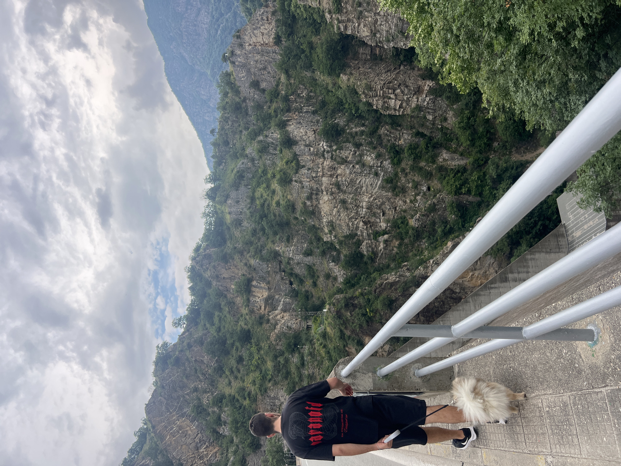 In Zhrebichko, Bulgaria, a man wearing a black t-shirt with red lettering and black shorts walks a small, fluffy dog on a leash along a paved walkway. The walkway is bordered by a metal railing and overlooks a steep, verdant gorge. Rocky cliffs covered in trees rise dramatically on the opposite side of the gorge. The sky is overcast with scattered clouds.