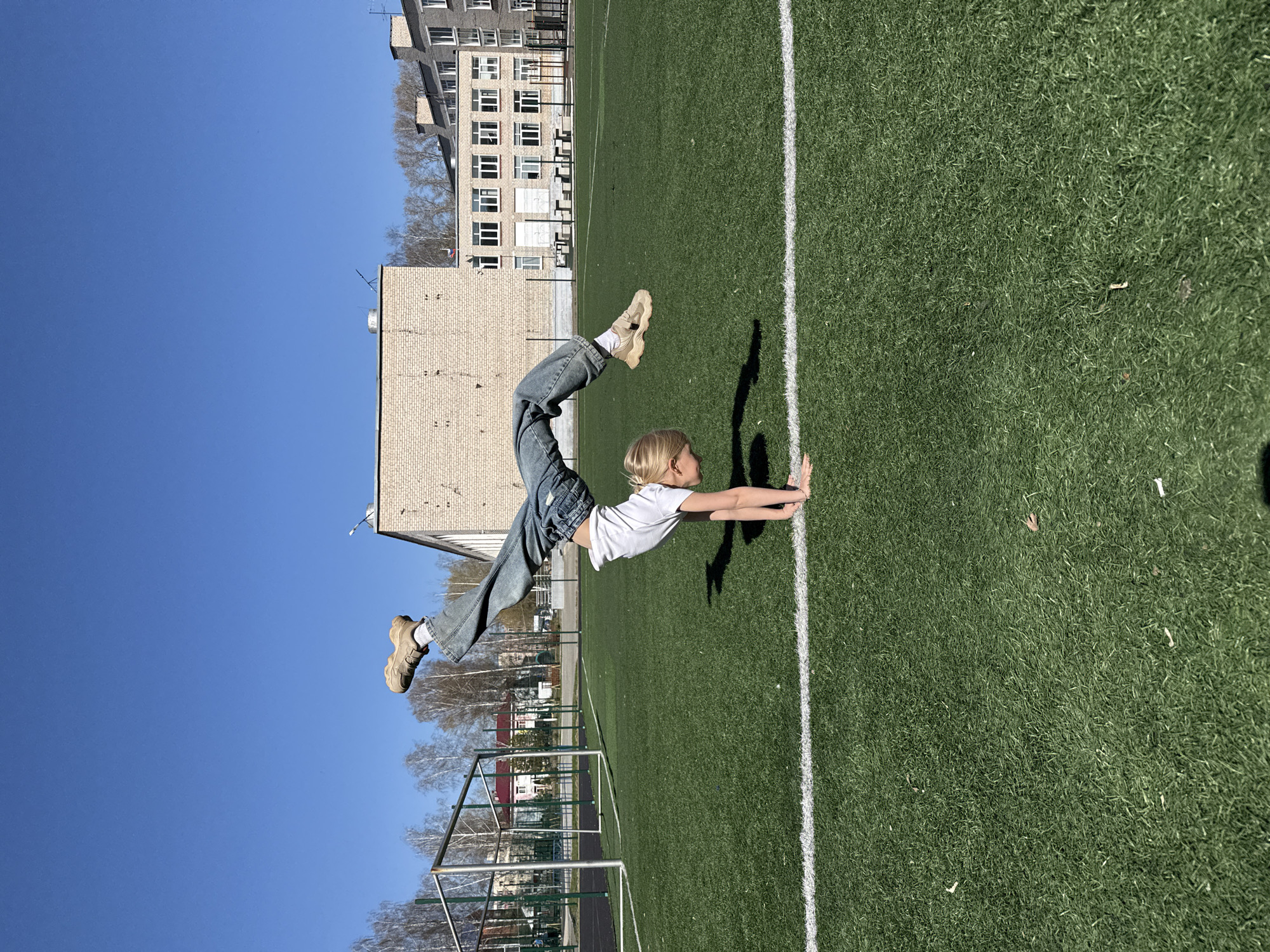 A young woman with blonde hair wearing a white T-shirt and blue jeans is performing a gymnastic move on a green artificial turf field in Chaikovsky, Russia. She is in a handstand position with her body arched. The background includes a building with several windows, possibly an apartment building, a clear blue sky, a soccer goalpost, and trees in the distance. The picture is taken in daylight with strong sunlight.