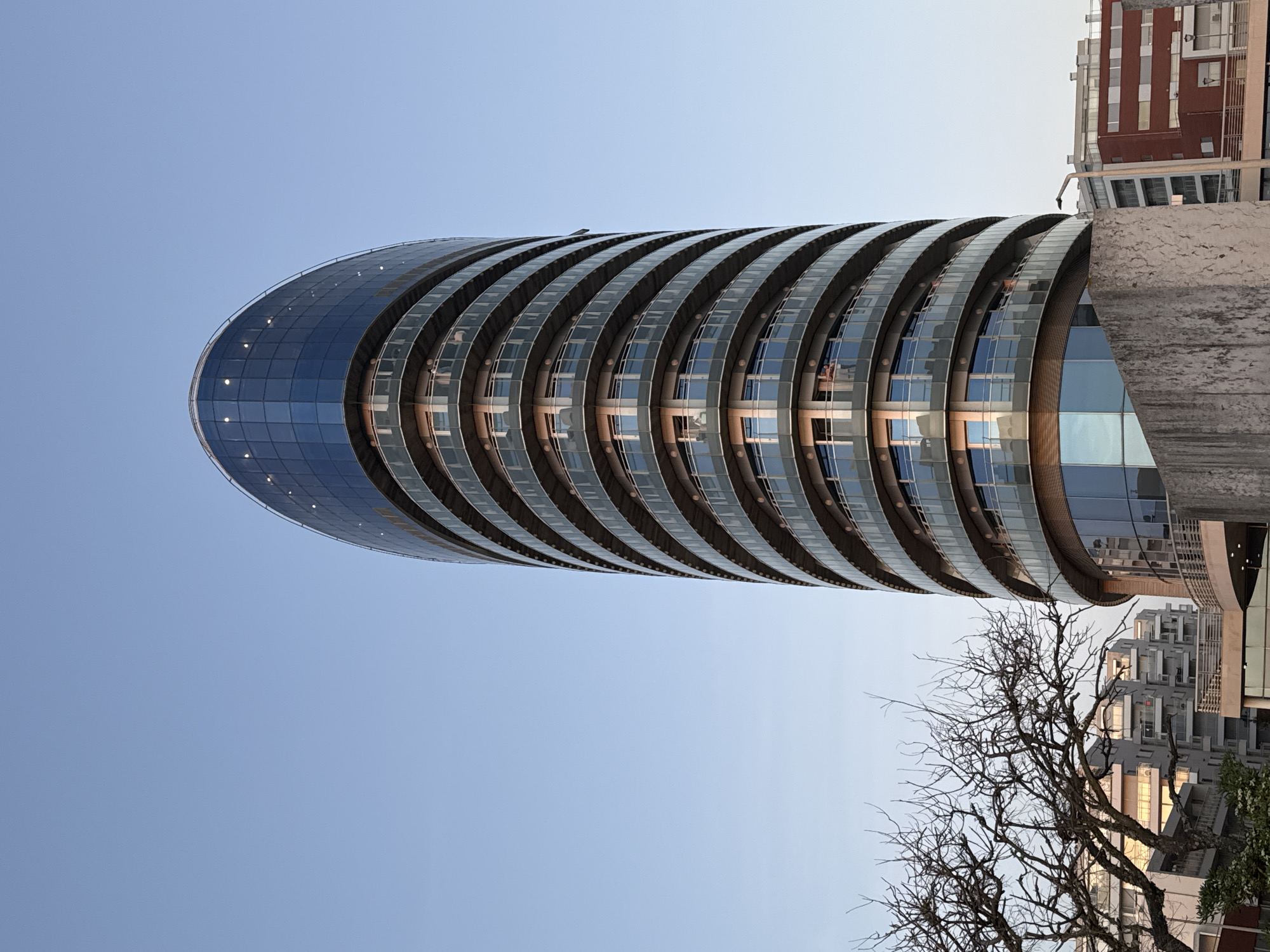 The image depicts a cylindrical, multi-story building with rounded ends. The building's façade is made of glass and has a layered, terraced design with what appear to be balconies encircling each level. The architecture is modern, with clean lines and a reflective surface. The sky behind the building is light blue. Parts of other buildings are visible to the right, including a brick-clad structure and a modern building with geometric cutouts, suggesting an urban environment. The bare branches of a tree are present in the lower right corner of the image. This building is located in the city of Figueira da Foz, Portugal.