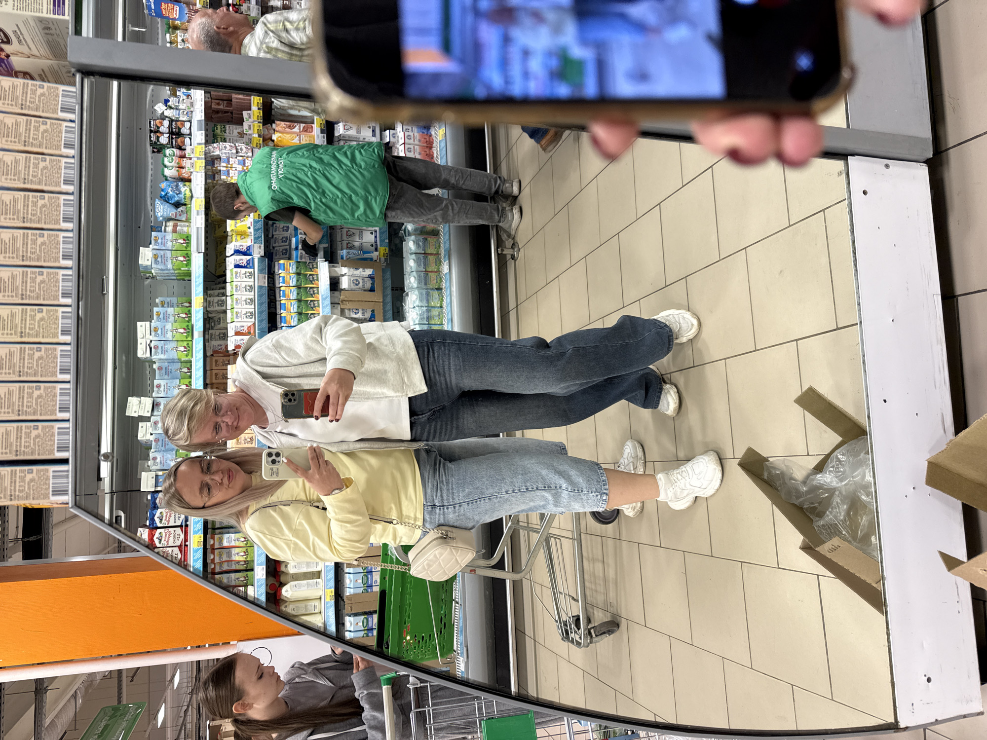 In an indoor marketplace, two women are captured in a mirror reflection. The woman on the left wears a yellow sweater, blue jeans, and white sneakers. She holds a white handbag. The woman on the right wears a white hoodie, blue jeans, and white sneakers. Both women are holding smartphones, seemingly taking photos. In the background, shelves are stocked with various products, and a shopping cart is visible. To the left of the women, a third person in a gray hooded jacket is partially visible. Further in the background, a man in a green vest is seen working, and another man in a plaid shirt is also present. The floor is tiled with a beige and light brown pattern. Cardboard boxes are visible in the foreground, on the floor.