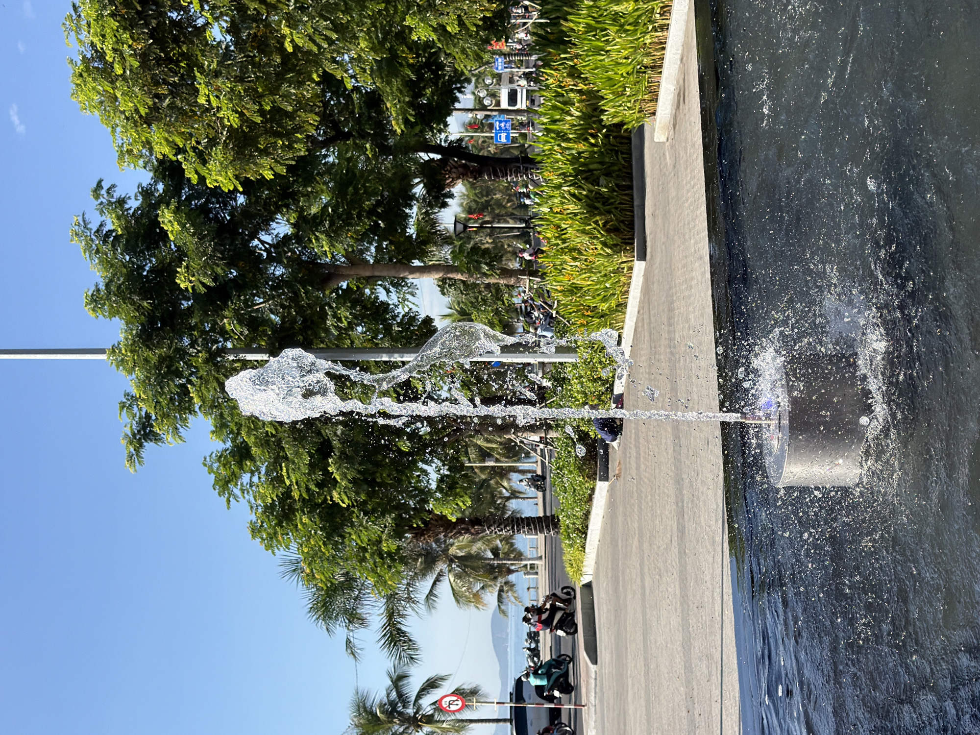 A shallow, dark water feature with a cylindrical metal fountain base is in the foreground, with water jets arcing upwards and splashing. Beyond the water feature, a paved walkway leads to a street lined with palm trees and lush green foliage. A road runs parallel to the beach, with several motorbikes carrying passengers. In the background, the sea is visible under a clear blue sky. The scene appears to be a public space in Nha Trang, Vietnam, likely a promenade or park area near the coast, given the presence of palm trees, the sea, and the tropical vegetation. The overall impression is one of a sunny, active urban environment.