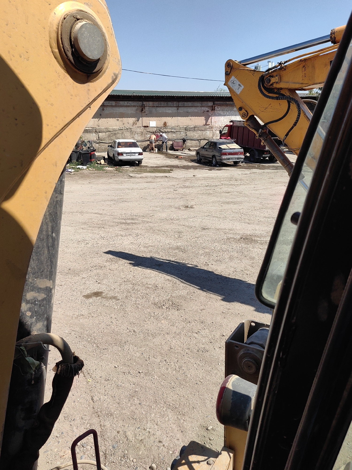The image depicts an outdoor industrial or construction area with a clear blue sky. In the foreground, parts of a yellow construction vehicle are visible, including a large circular metal component and a dark hose with a frayed end. The ground is composed of gravel and dirt, with some scattered debris and a dark stain.
In the middle ground, two cars are parked on the gravel. The car on the left is white with a license plate that reads "E245CP 71". The car on the right is darker and appears to be a sedan, also with a visible license plate. Behind the cars, a red truck with a dark cargo bed is parked.
Further back, a low concrete wall runs horizontally across the scene, topped with a corrugated metal structure that appears to be a roof or awning. Three individuals are present near the white car, two of whom appear to be engaged in washing something. One person is wearing a light-colored shirt and shorts, and another is wearing a dark shirt and light-colored pants.
The overall impression is a functional outdoor space, possibly a service yard or construction site, in Shymkent, Kazakhstan, characterized by heavy machinery, vehicles, and a utilitarian building.