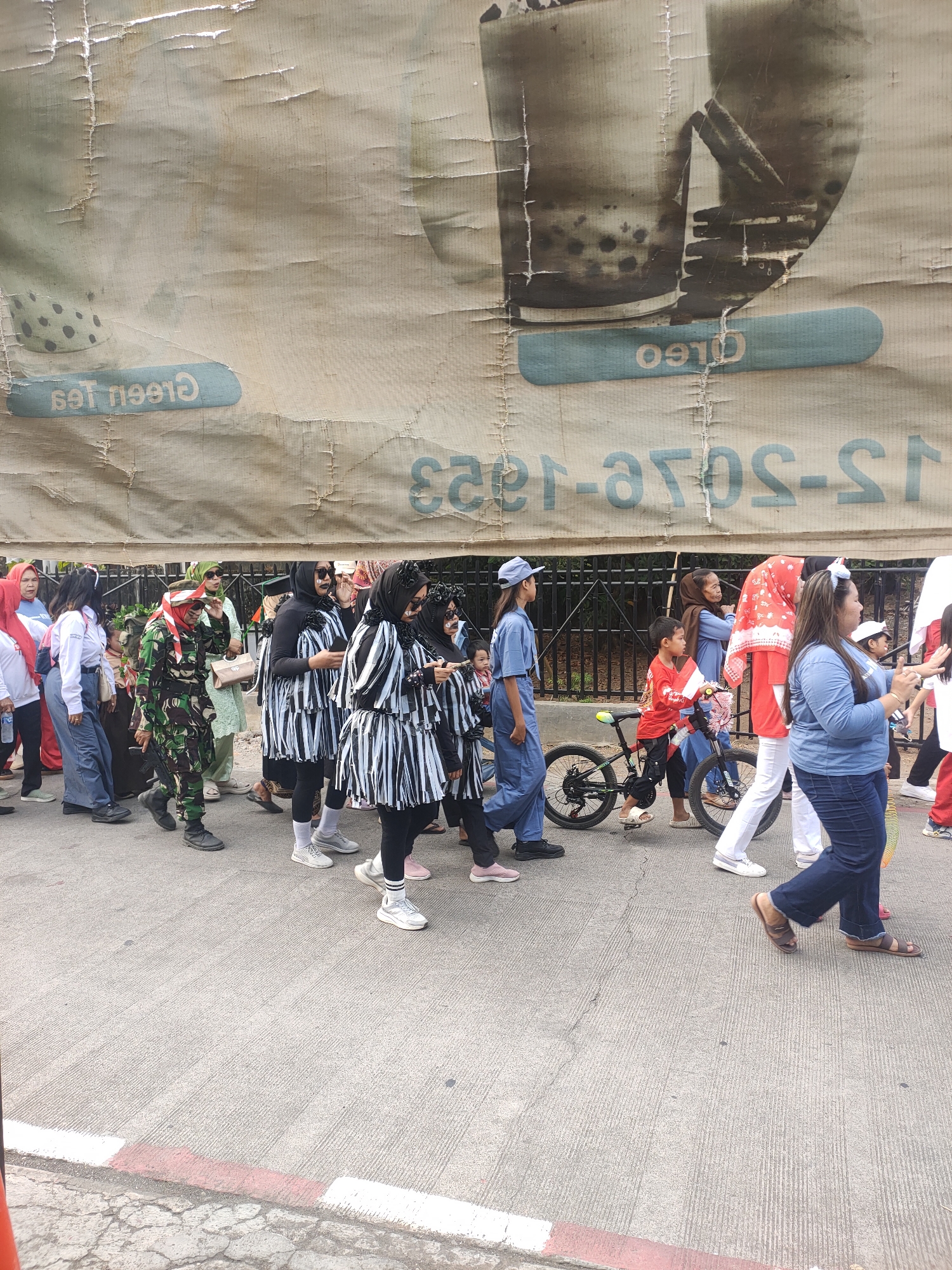 In Subang, Indonesia, a group of people are participating in a parade along a paved street, likely celebrating a local event or holiday. The scene is dominated by a large, faded banner overhead, featuring images of drinks and text that appears to be advertising. The banner has visible wear and tear, suggesting it has been exposed to the elements for some time.
The parade participants display a variety of attire and activities. Towards the left, a person in a camouflage military uniform and a red and white headscarf stands out. They are holding what appears to be a prop rifle. To their right, a group of women are dressed in matching black and white striped outfits and headscarves, with some having dramatic face paint resembling skulls. Further down the line, a young boy rides a bicycle decorated with Indonesian flags, accompanied by adults. The crowd includes individuals of different ages, many wearing casual clothing, jeans, and sneakers. The street is bordered by a black metal fence and some foliage, hinting at a public space or a residential area. The pavement shows signs of age, with cracks and distinct markings at the edge. The overall atmosphere suggests a community gathering, with a mix of patriotic and festive elements.