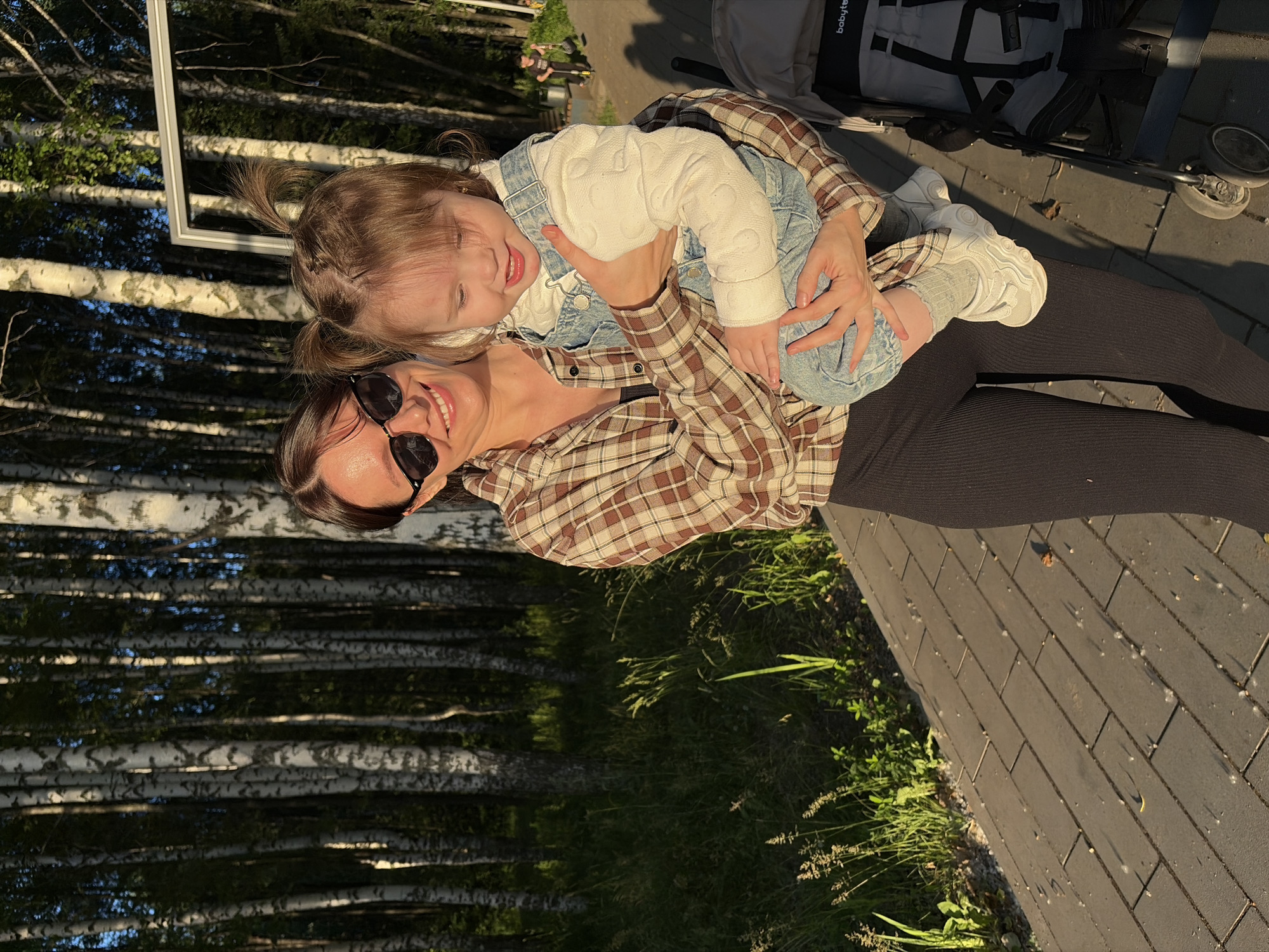 In the city of Chaikovsky, Russia, a woman wearing a plaid shirt and black leggings holds a baby wearing a denim romper and a white long-sleeved shirt. The woman is wearing sunglasses and smiling at the baby. The baby is looking down and smiling. They are standing on a paved pathway next to a stroller. In the background, there is a forest of birch trees. The sun is setting, casting long shadows.