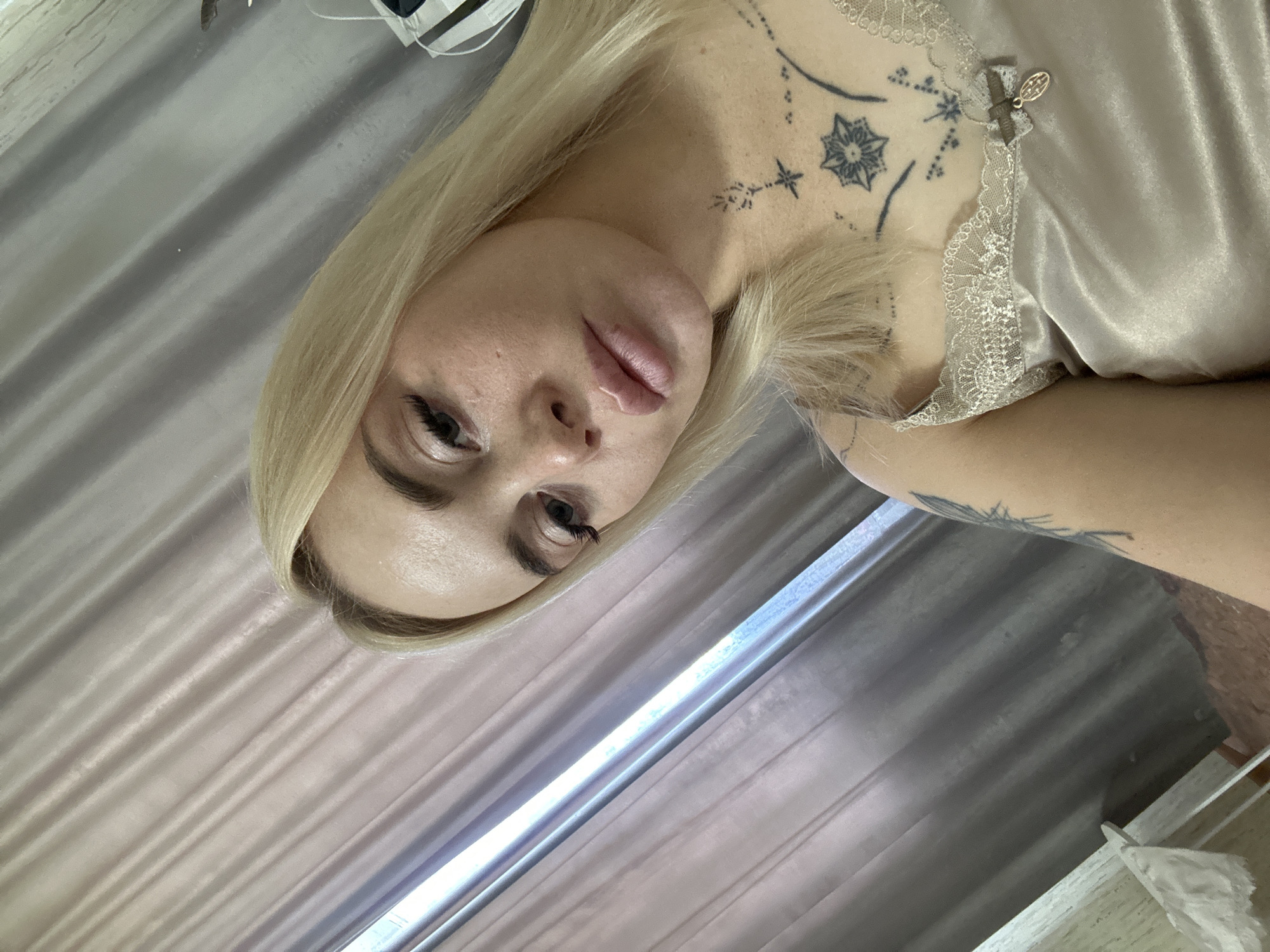 A young woman with blonde hair and tattoos is posing for a selfie indoors. She is wearing a light-colored satin camisole with lace trim. The background consists of grey curtains and a window with a blue light shining through.