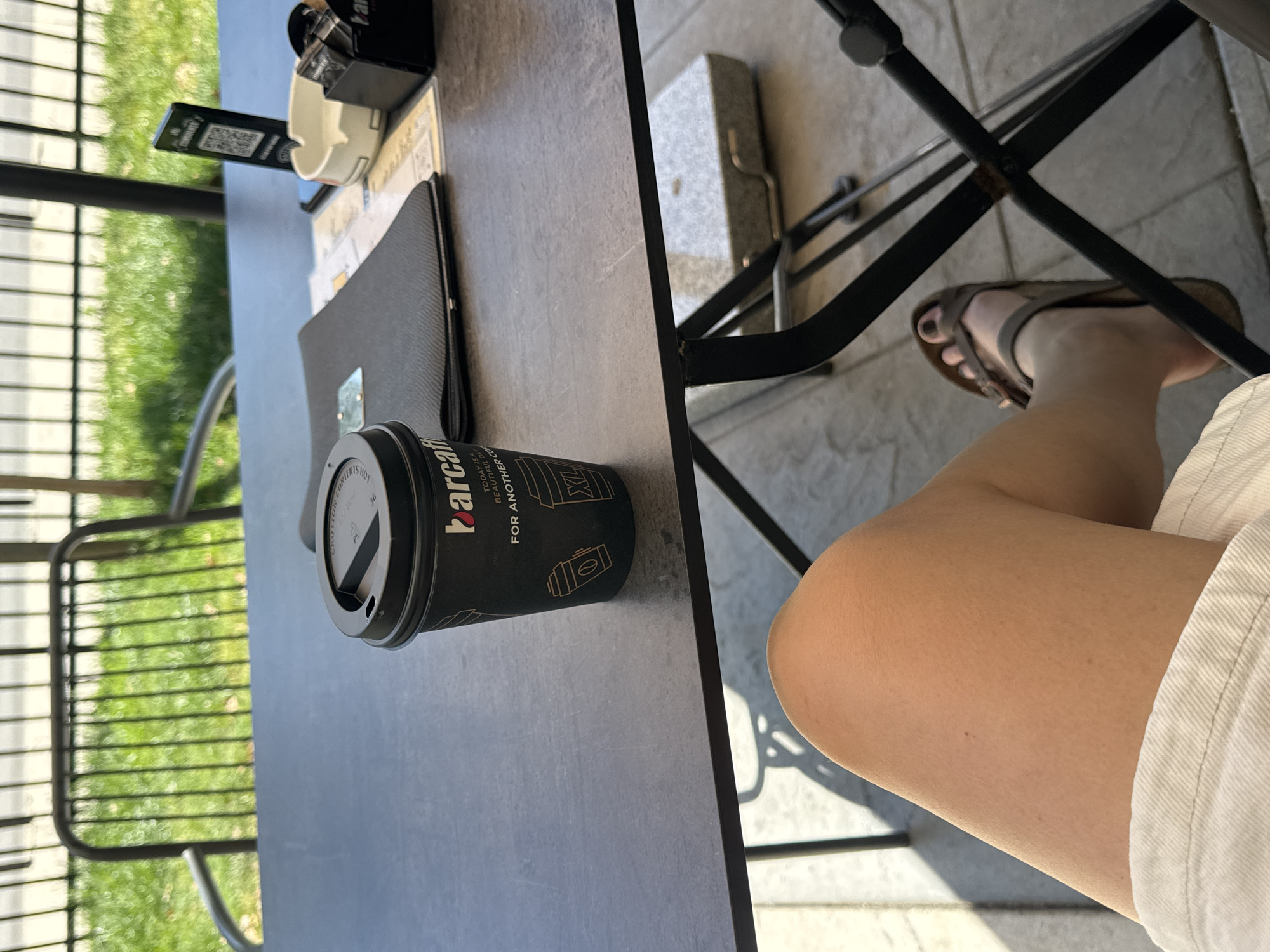 A black paper cup with a black lid bearing the brand "barcaffe" is placed on a grey table next to a black menu and a receipt holder. The table appears to be outdoors. Adjacent to the table is a black metal chair. To the right of the table and chair is a person's leg clad in white shorts, with a foot visible in a sandal. A chair and green foliage are visible in the background. Based on the brand of coffee cup, this image was likely taken in Podgorica, Montenegro.