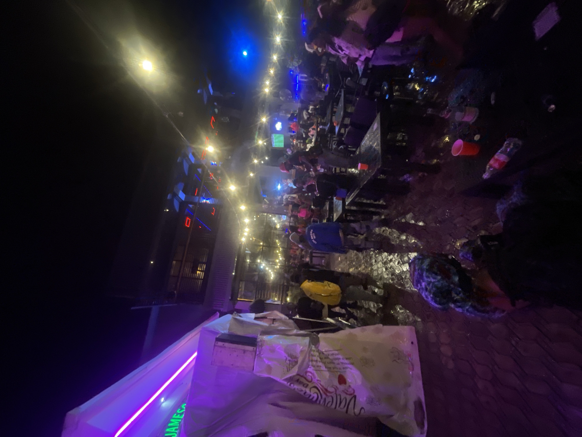 The image shows an outdoor bar or restaurant in Lokoja, Nigeria, at night. The area is illuminated by string lights and LED lighting. Multiple people are seated at tables, with some standing and interacting. There are empty cups and bottles on the tables. A Valentine's Day themed bag and other items are placed on a bar area with illuminated neon lettering. A building with visible red accents stands on the left side.