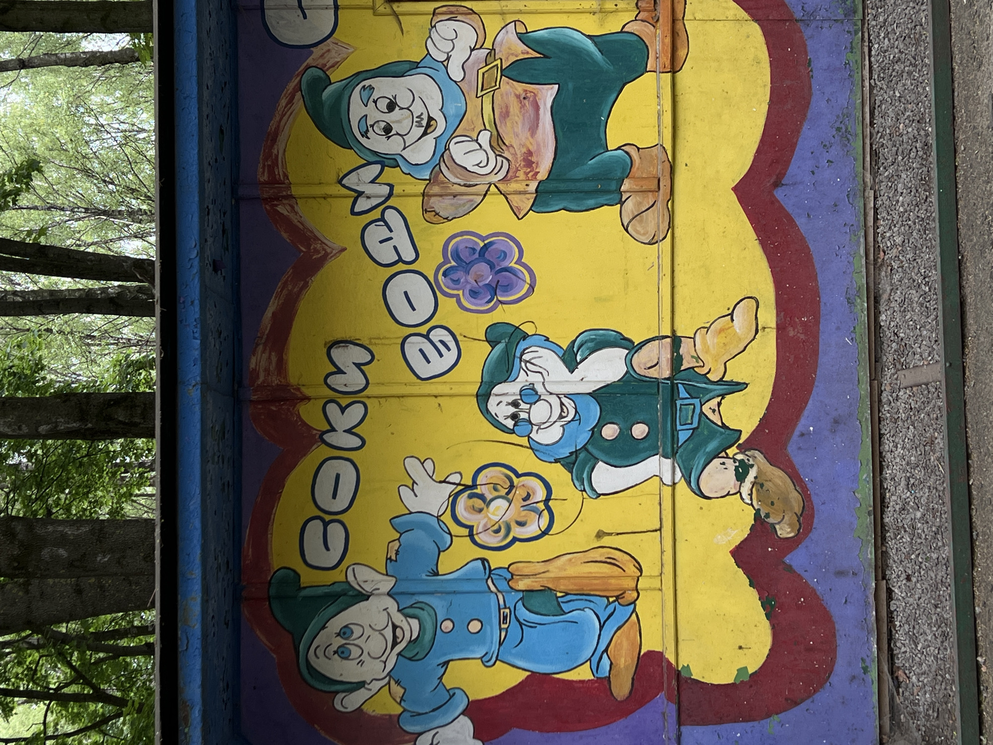 The image shows a section of a painted structure, likely a wall or barrier, featuring cartoon depictions of gnomes in a whimsical style. The gnomes are painted in various shades of blue and green, with yellow and purple flower accents around them. In large white letters is the name "ВОВЧКИВ". The artwork is rendered on a yellow background bordered by a scalloped edge in red and purple. The painting exhibits signs of age and wear, with areas of peeling paint and discoloration. The structure is located in the city of Ternopil, Ukraine. Adjacent to the artwork on the left is a glimpse of trees and foliage, while on the right, a section of a paved walkway is visible.