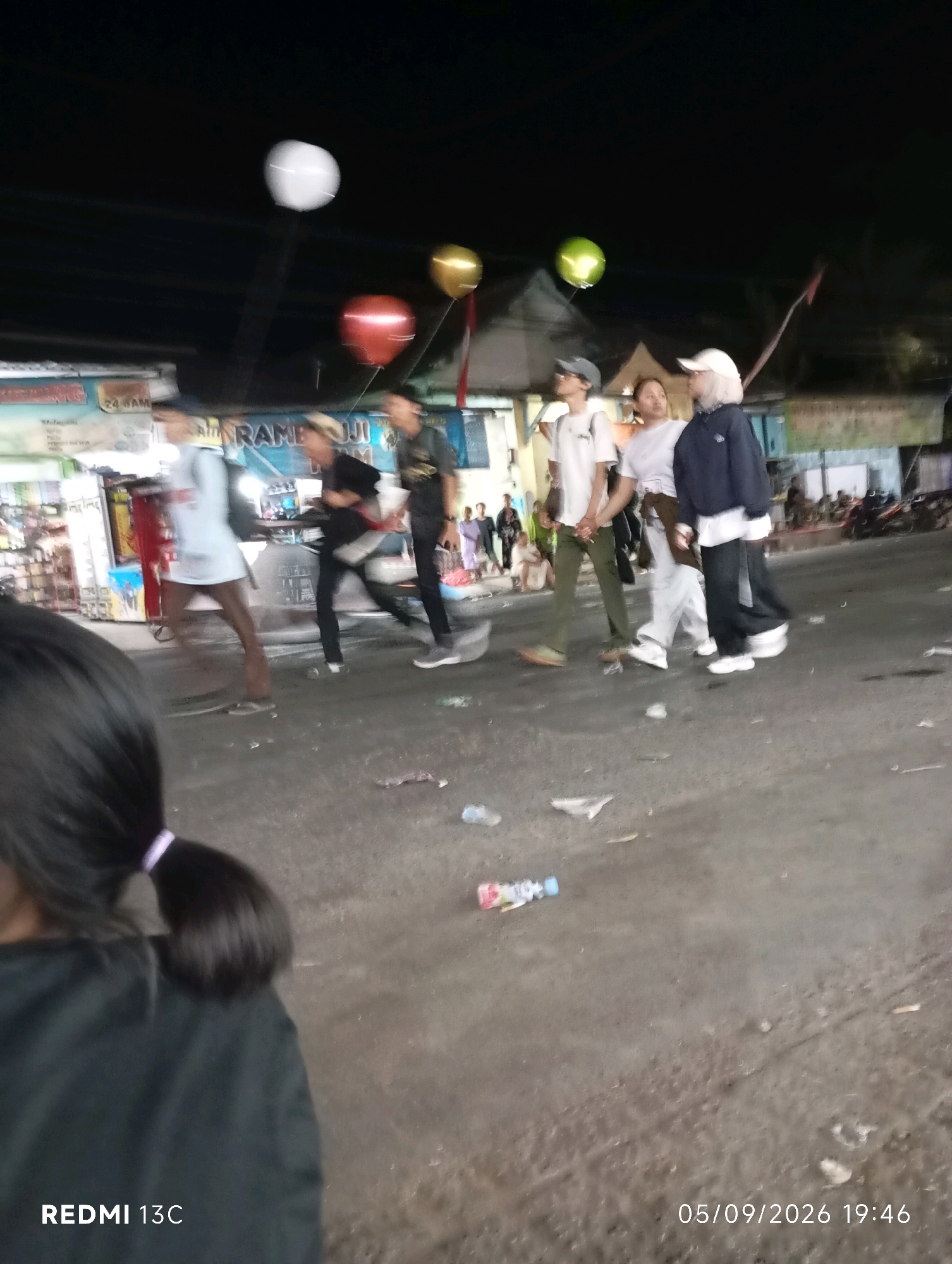 The image, timestamped 05/09/2026 19:46, captures a street scene at night in Rambipuji, Indonesia. In the foreground, the back of a person's head with dark hair tied in a ponytail with a light purple elastic band is visible, wearing a black garment. The middle ground displays a group of people walking and standing. Among them, three individuals are prominent: one person in a light-colored long-sleeved shirt and brown pants, another in a dark t-shirt and black pants, and a third in a dark jacket over a white shirt and dark pants, wearing a white cap. Several people are also seen in the background, some gathered near stalls or buildings. The environment appears to be a street with a paved surface, littered with debris, including plastic bottles and paper. In the upper portion of the image, three balloons are visible: one white, one red, and one yellow. Behind the people, buildings with illuminated signs and structures suggest a commercial or public area. The overall lighting indicates it is nighttime.