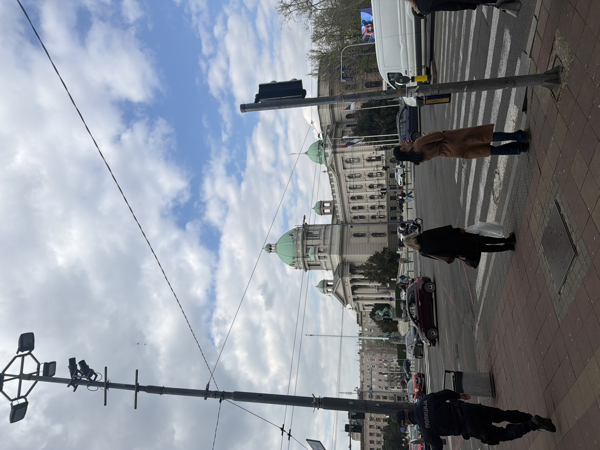 The image shows a street scene in Belgrade, Serbia, with the Serbian Parliament building in the background. The sky is cloudy with patches of blue. A traffic light pole stands in the foreground. Pedestrians are crossing the street at a zebra crossing. Several cars are parked or driving on the street. A police officer in uniform is walking on the sidewalk. The sidewalk is paved with red bricks. A white van is parked near the parliament building. Flags are displayed on the building.