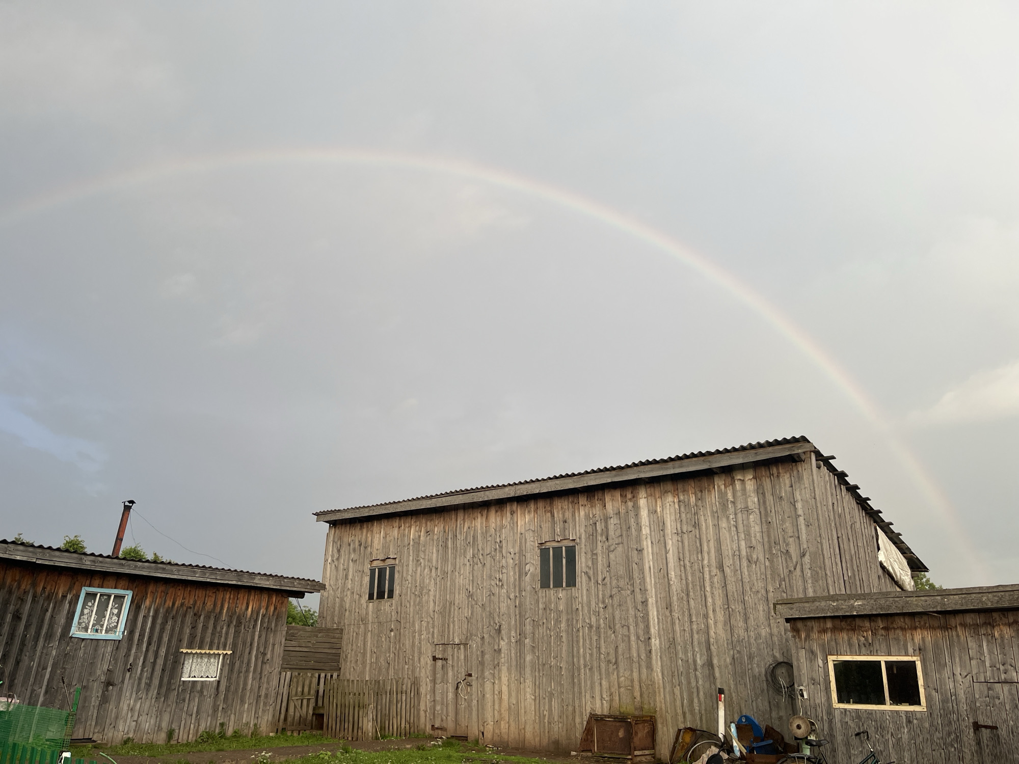 The image shows a rural scene with a prominent rainbow arching across a cloudy sky. In the foreground are two wooden buildings, likely sheds or outbuildings, constructed from vertical wooden planks. The larger building on the right has a corrugated metal roof and several dark rectangular windows. The smaller building on the left also has a corrugated roof and a distinct blue-trimmed window with white patterned curtains, and a smaller window above it. A metal pipe or chimney extends from the roof of the left building. A wooden fence, some of which appears dilapidated, separates the buildings from a grassy area. Various objects are visible near the buildings, including what appears to be old metal containers, a bicycle, and a green wire fence or bin in the lower left corner. The overall impression is one of a utilitarian, possibly aged, rural property under a sky that has recently experienced rain, as indicated by the rainbow.