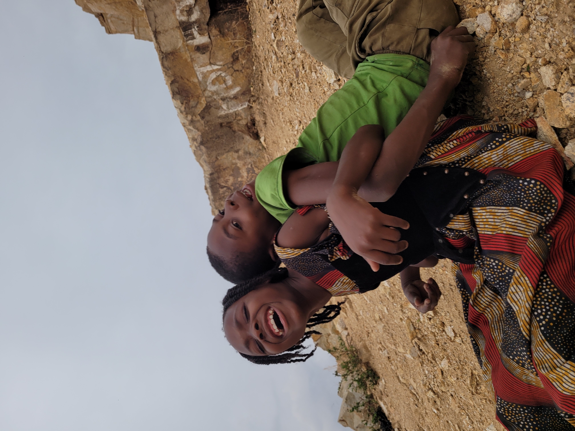 Two children are photographed against a backdrop of arid, rocky terrain, possibly a quarry or hillside. The child in the foreground is a girl, laughing with her mouth open, wearing a colorful patterned dress with a black bodice. The child behind her is a boy with a green shirt and olive green trousers. Their arms are around each other, suggesting an embrace. The setting appears to be in Barakin Akawo, Nigeria, based on available information. The sky is overcast.