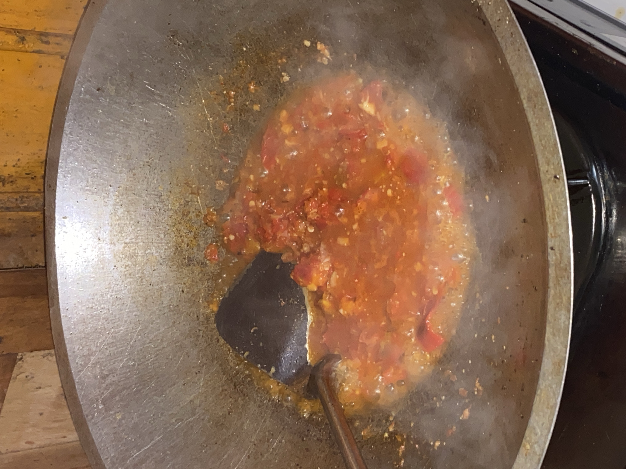 In a wok, a red sauce with visible chunks of chili and garlic is being stirred with a dark spatula. The wok is made of a dull metal and shows signs of wear and past use. Steam rises from the sauce, indicating it is actively cooking. The background consists of wooden planks, suggesting a kitchen environment. The scene appears to be in a home kitchen located in Ta', Indonesia, where a meal is being prepared.