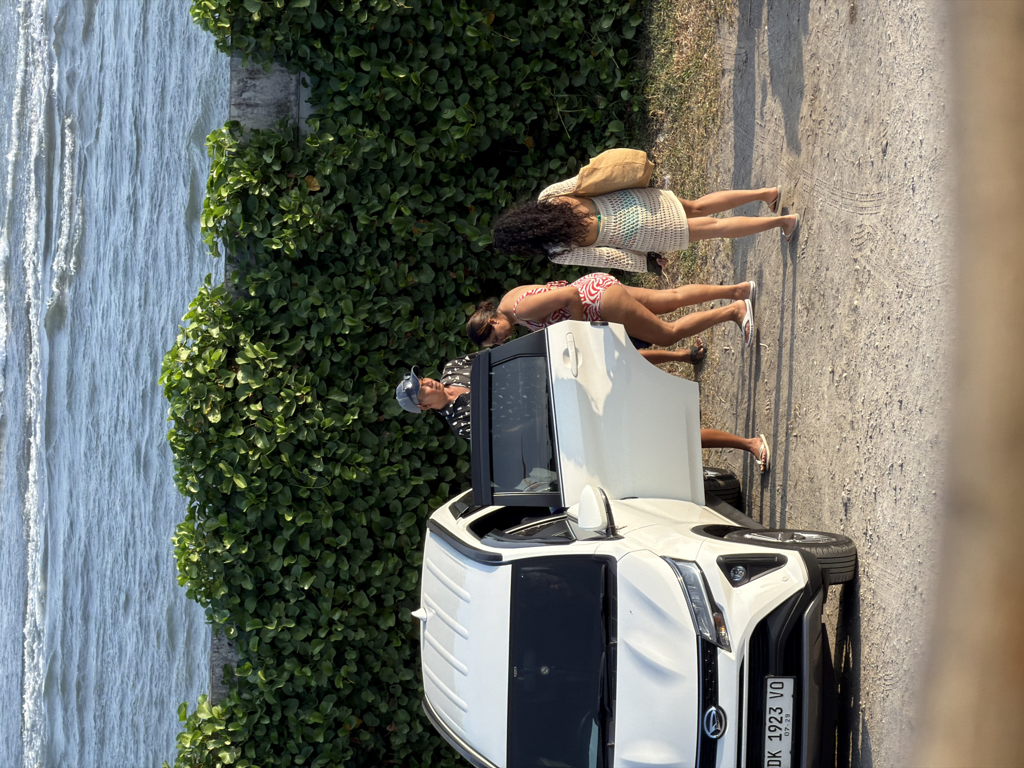 In the city of Mengwi, Indonesia, a white SUV with its driver-side door open is parked on a dirt road. Three individuals stand near the open car. One person, wearing a patterned shirt and a baseball cap, stands by the driver's side door. Another person, in a red and white striped swimsuit, stands to the right of the first individual, facing away from the camera. The third person, wearing a white crochet dress over a bikini and carrying a beige tote bag, stands to the right of the second person, also facing away from the camera. Behind the car and people is a dense green hedge, and beyond that, the ocean with gentle waves. The ground is covered in dirt and some patches of dry grass. The vehicle's license plate reads "DK 1923 VO".