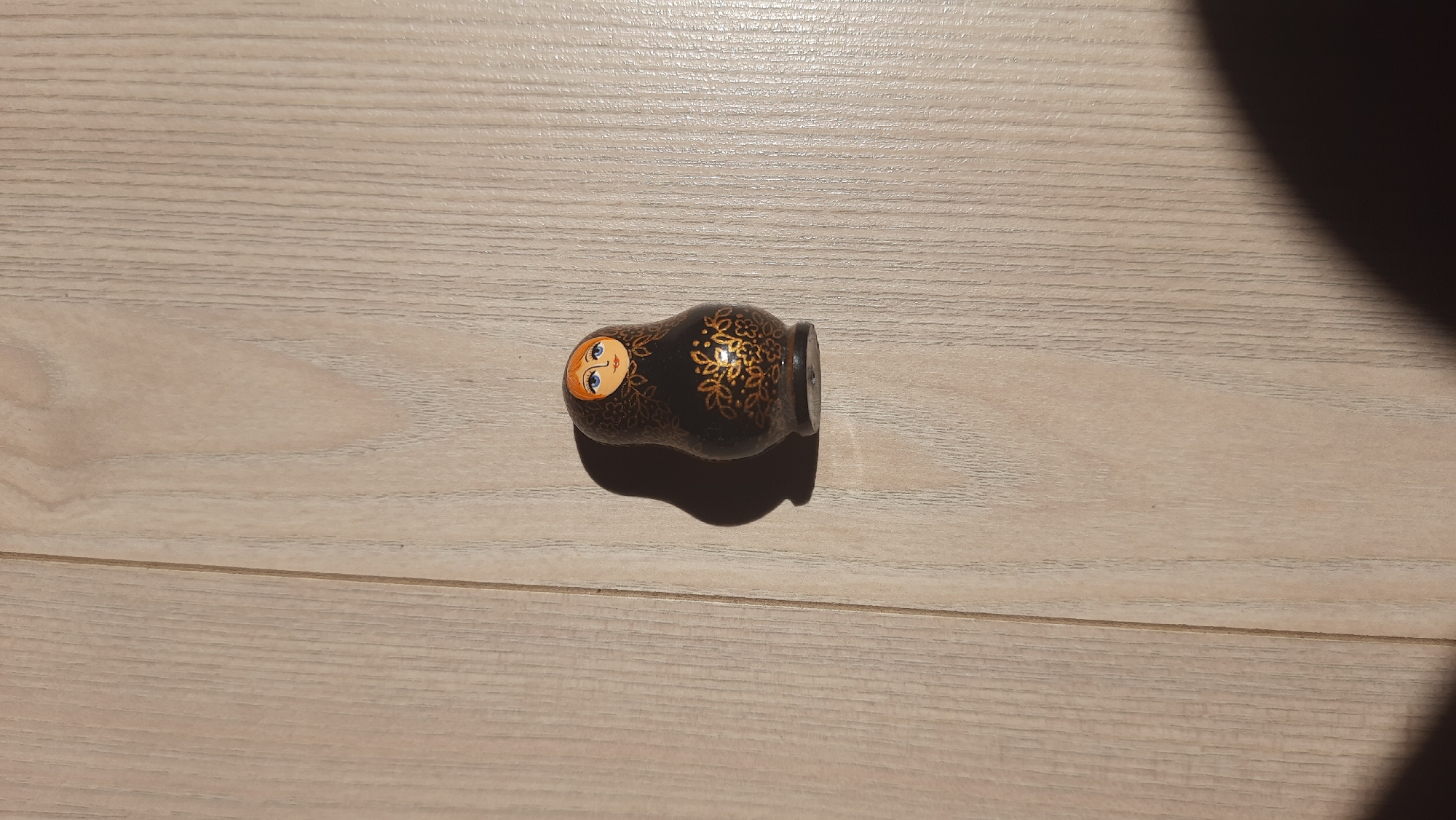 A single, small, black Matryoshka doll with gold floral patterns and a painted face with blue eyes and orange hair is positioned vertically on a light brown wooden floor. The doll is casting a dark shadow to its left. The floorboards are laid horizontally in the image and have a visible grain. The image is an overhead shot. There is no discernible city or location information.