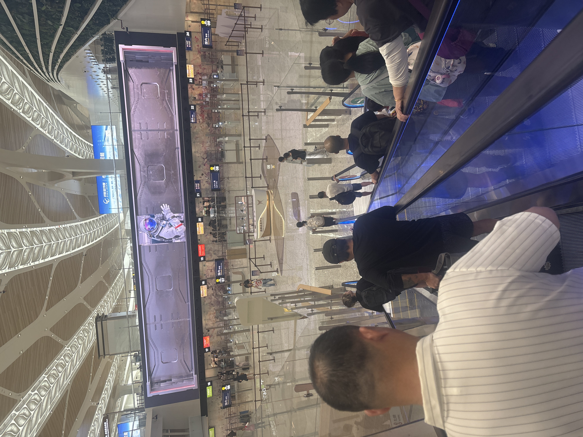 In the city of Baiyun District, China, a modern airport terminal features a wide, well-lit escalator with blue LED lights. Several individuals are ascending the escalator, their backs to the camera. In the foreground, a man wearing a white and grey striped polo shirt is visible. Behind him, other passengers are seen, including a man in a black t-shirt and shorts and a woman in a light green jacket. The escalator passes over a polished floor with reflective surfaces. In the background, beyond the escalator's railing, is a spacious, modern airport concourse. Numerous transparent barriers and stanchions delineate various queues and security checkpoints, with signs in Chinese characters visible. A large digital display screen hangs high above the concourse, showing an image of an astronaut and what appears to be a spacecraft interior. People are milling about the concourse, some queuing at the checkpoints. The ceiling of the terminal is a complex, architectural design with illuminated panels. The overall scene depicts a busy, contemporary airport environment.
