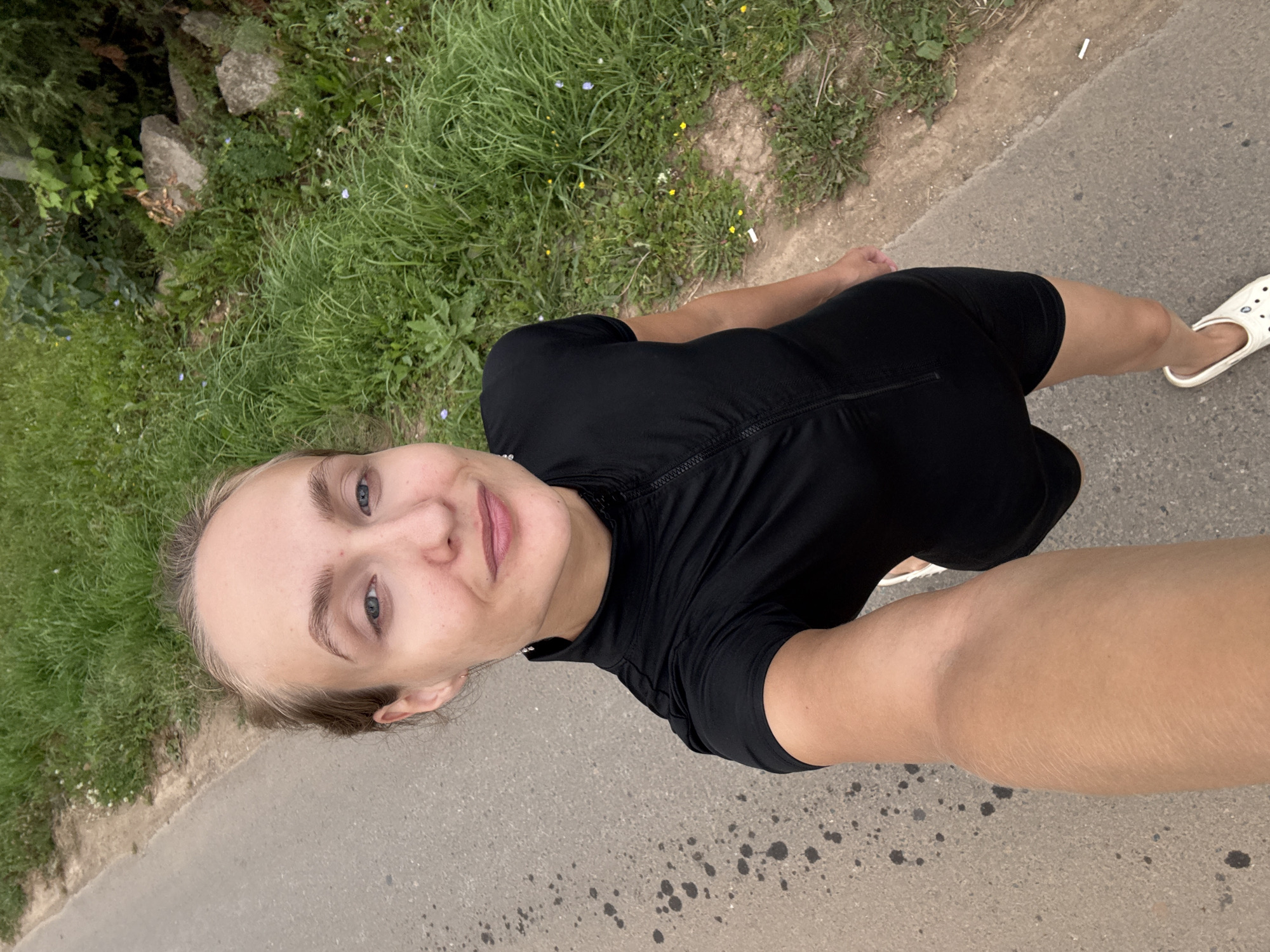 A woman stands on a paved path, holding a camera in her left hand to take a selfie. She is wearing a black short-sleeved zippered top and black shorts. Her hair is pulled back. The path is bordered on the left by a grassy area and on the right by more grass and some rocks. The location is specified as Podshivalovo, Russia.