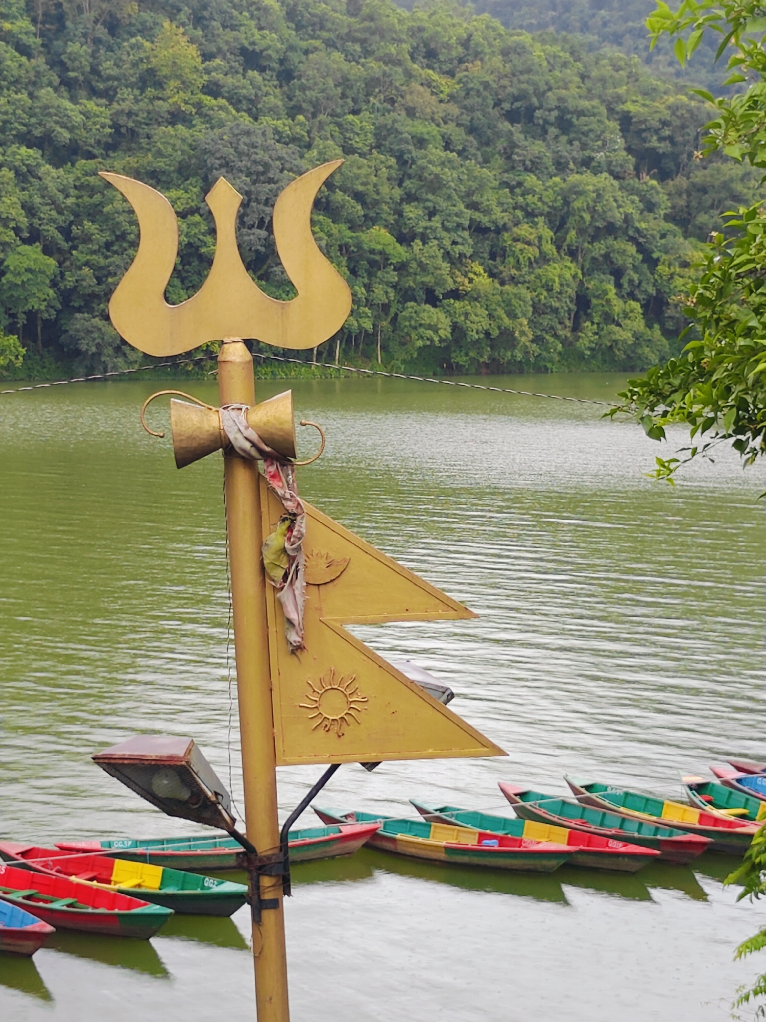 In Pokhara, Nepal, a golden trident-like religious symbol stands prominently on the shore of a green lake. The trident is adorned with colorful fabric tied around its central pole. Below the trident, a yellow pennant with a sun symbol is attached. A streetlamp with a rusted metal casing is mounted on the pole, facing the water. Several brightly colored boats, including red, yellow, and green, are moored along the edge of the lake. In the background, a lush green hillside densely covered with trees slopes down towards the water, indicating a natural, possibly mountainous, landscape. The water is calm with small ripples, suggesting a gentle breeze or perhaps light rain. The scene captures a tranquil moment by the lake, combining religious iconography with recreational elements.