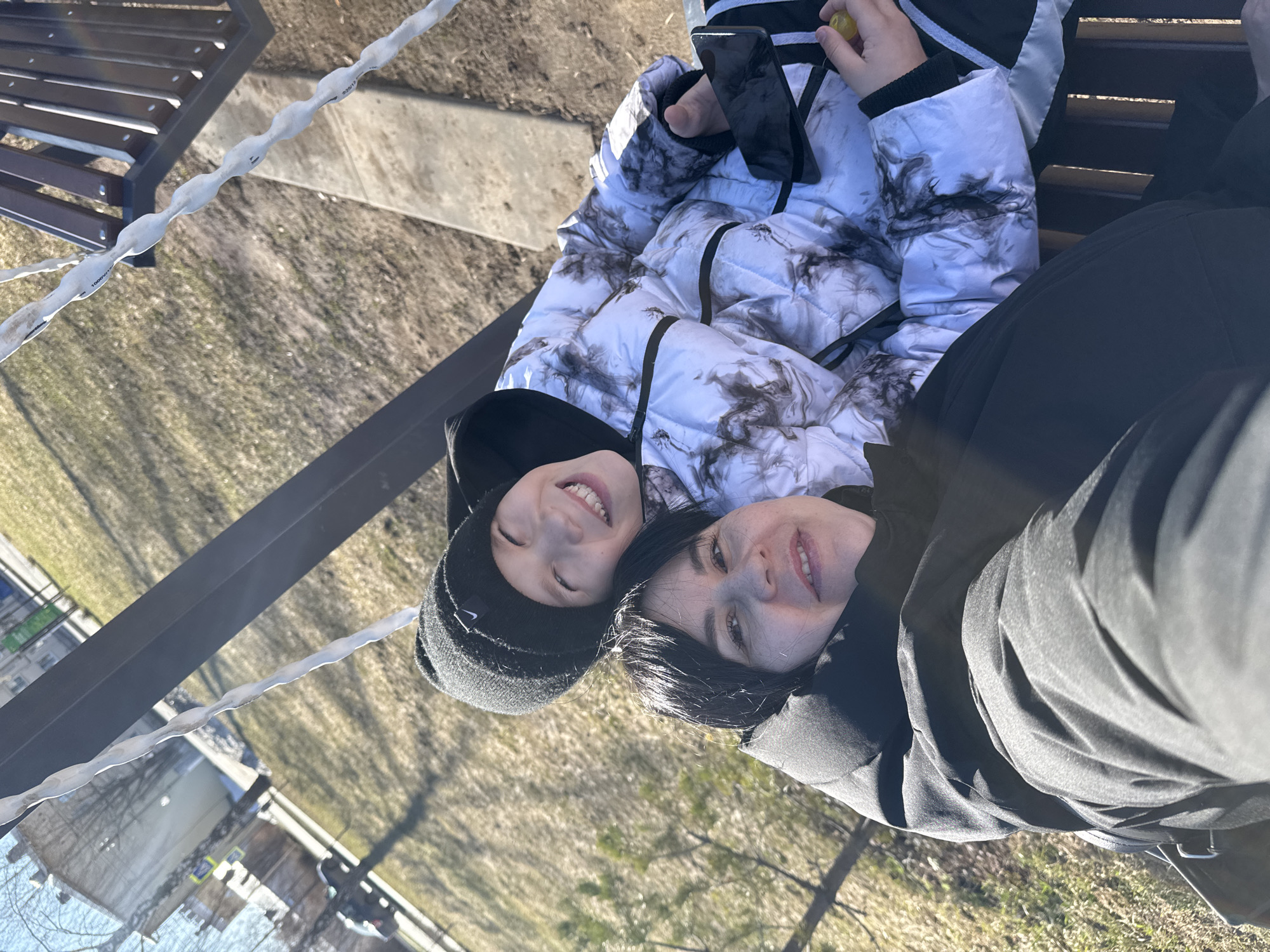 A photo shows a woman and child lying together on a bench. The child is wearing a black beanie and a white jacket with gray and black accents. They are holding a phone in their right hand and a yellow object in their left. The woman is wearing a black jacket and has black hair. Both are smiling. The bench has a metal frame and wooden slats. The ground is covered in dry grass and some buildings are visible in the background. The location is possibly in Zav'yalovo, Russia.