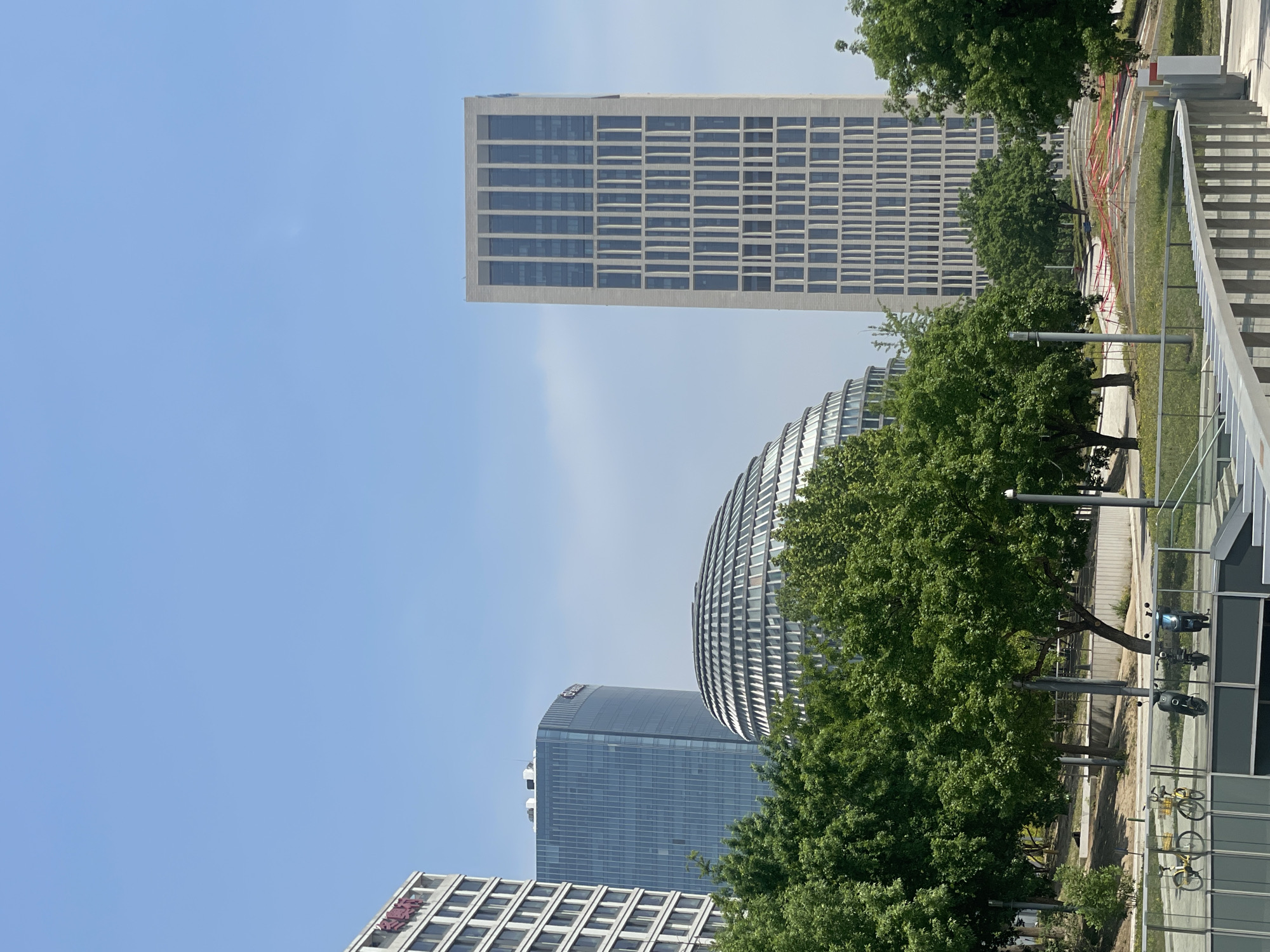 The image captures a view of buildings in Chaoyang District, China. Specifically, the Beijing Ginza Mall, a cylindrical building with a visible grid pattern exterior, is seen. The building is surrounded by green trees. Alongside it is a tall rectangular building with a similar grid pattern facade. In the foreground are more trees and a glimpse of a modern structure with a reflective surface, as well as bicycles parked. The sky is clear and blue.