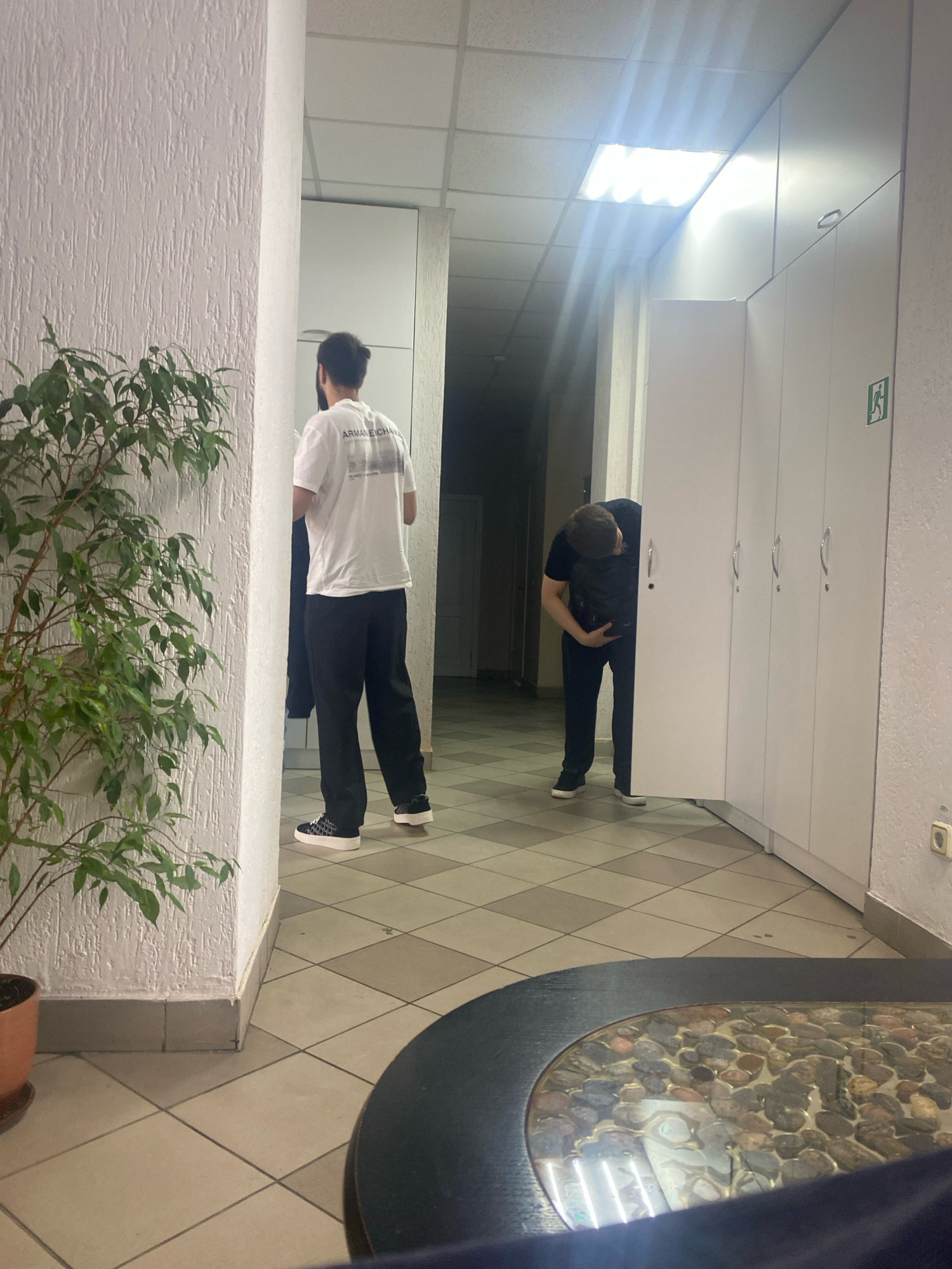 The image depicts an indoor hallway or common area with a white textured wall on the left, a large potted plant, and a tiled floor. In the foreground, a dark, circular object, possibly a table, with a reflective surface containing stones is visible.
Further down the hallway, on the right, there is a row of white lockers with silver handles. A person wearing a black t-shirt and dark pants, with a black backpack, is bent over, partially obscured by an open locker door, appearing to be interacting with it. To the left of this individual, another person is standing, facing away from the camera, wearing a white t-shirt with a graphic print and black pants. They are standing near a doorway.
The environment appears to be a public space, possibly a locker room, office break room, or a similar facility. There is a fluorescent light fixture on the ceiling, casting a bright light. An exit sign with a green pictogram is visible on the wall near the lockers. The overall impression is a mundane, everyday scene.
It is not possible to determine the exact location in Minsk, Belarus, from the visual information provided in the image.