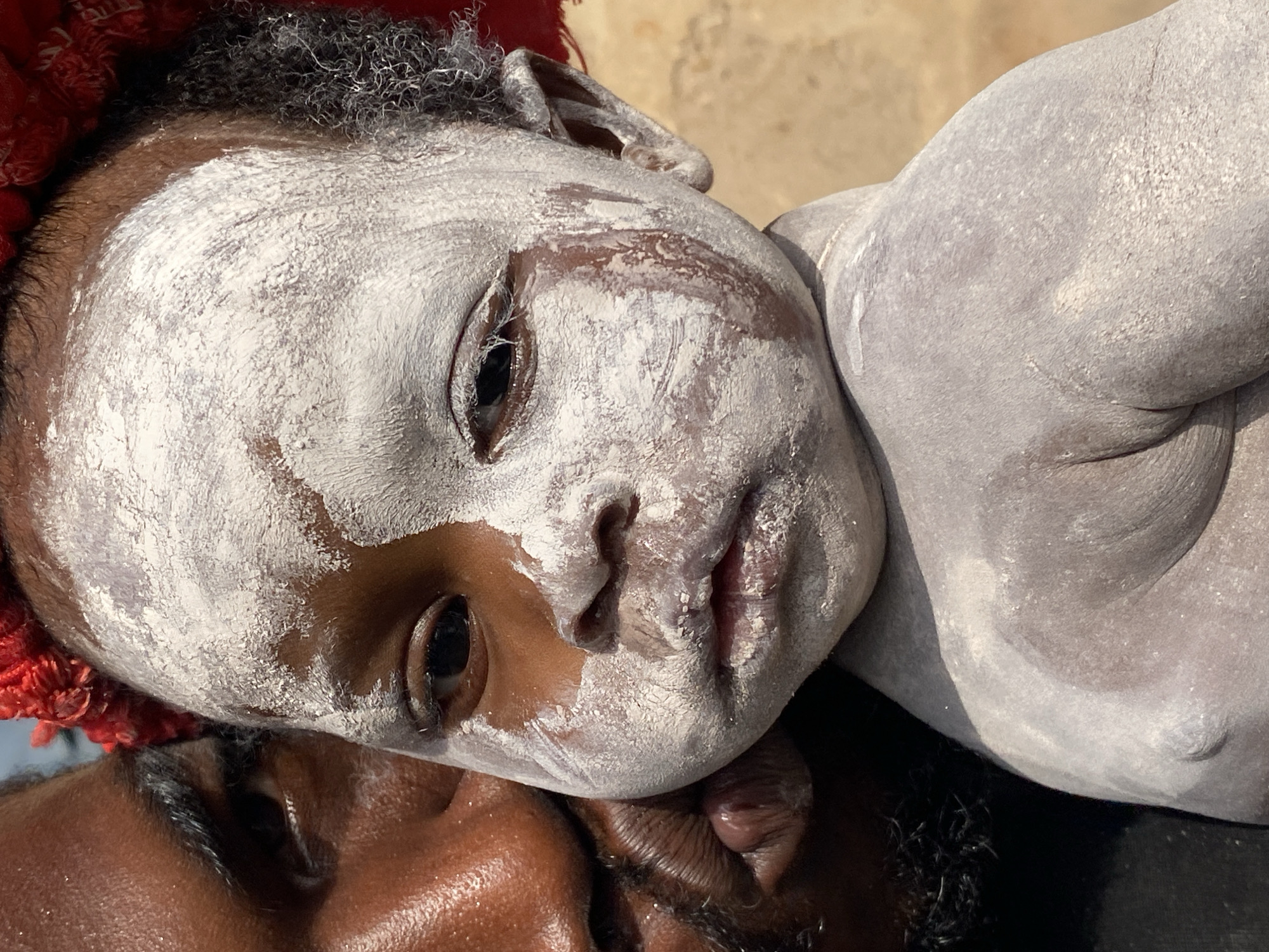 This is a close-up, eye-level shot of a dark-skinned person holding a baby. The baby is partially covered in white powder, with a streak of it running down its face. The baby's left eye is open, looking towards the viewer, while its right eye is partially closed. The person holding the baby has dark skin and a beard, and is leaning in towards the baby. A red object, possibly a headdress or decoration, is visible at the top of the frame. The background is out of focus and appears to be an outdoor environment with a muted brown color. The image was taken in Ufu Esioni, Nigeria.