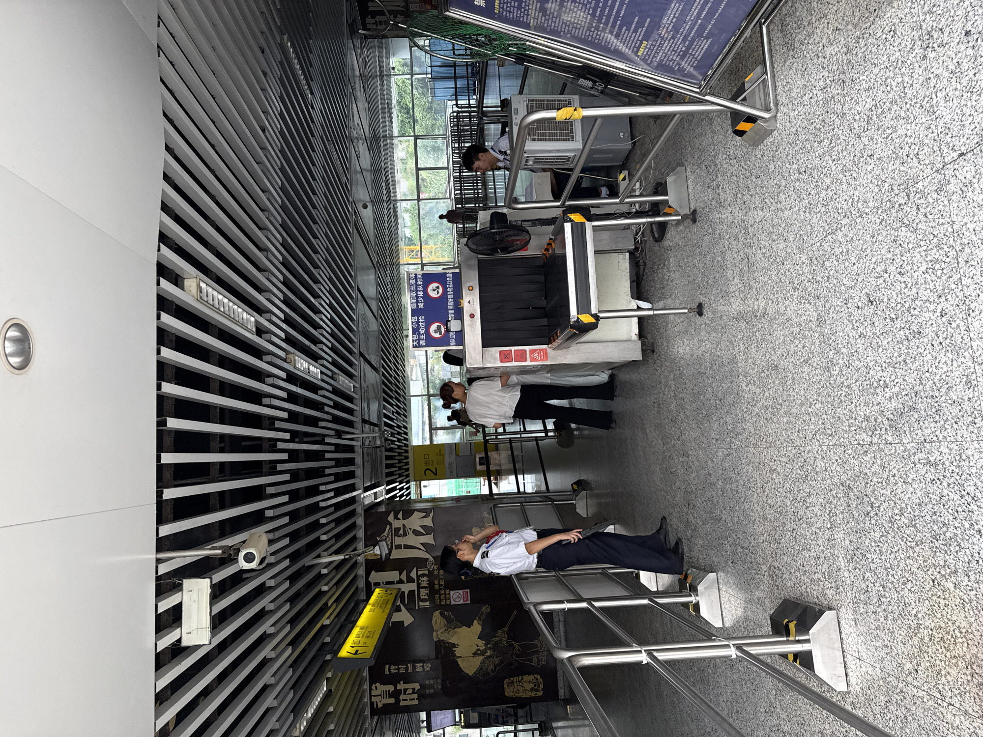 In the city of Shapingba District, China, at a security checkpoint, two security personnel are visible. One individual, wearing a white shirt and dark trousers, stands near a black and yellow stanchion, holding a dark object. Another security officer in a white shirt and black trousers is seated near an X-ray scanner, with a fan and an air conditioning unit beside them. Several individuals are queueing to pass through the security scanner. The environment includes a metal ceiling with light fixtures, metal railings, a speckled grey floor, and large signs with Chinese text. The scene suggests a transportation hub or public facility undergoing security screening.