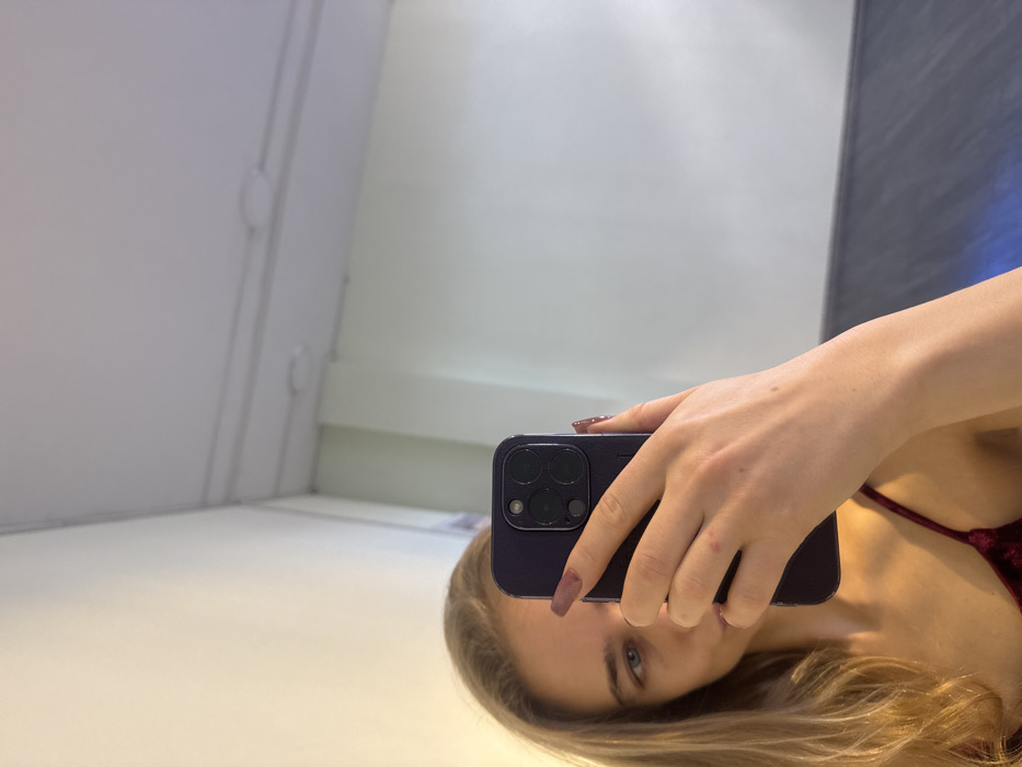 The image depicts a mirror selfie of a female individual taken indoors. The woman, with blonde hair and light-colored eyes, is visible from the mid-chest upward. She is wearing a dark red or burgundy velvet spaghetti strap top. Her right hand holds a dark-colored smartphone, an iPhone model identifiable by its prominent three-camera array, obscuring the lower half of her face. Her fingernails are manicured with a reddish-brown polish. The reflection in the mirror shows a room with light cream or off-white walls and a white ceiling. The ceiling features visible white electrical conduits running horizontally and two circular surface-mounted light fixtures. A dark blue or grey surface is visible in the lower right portion of the reflected background, possibly furniture or a lower wall section. The exact location within a city of Unknown, Unknown cannot be determined from the provided visual information.