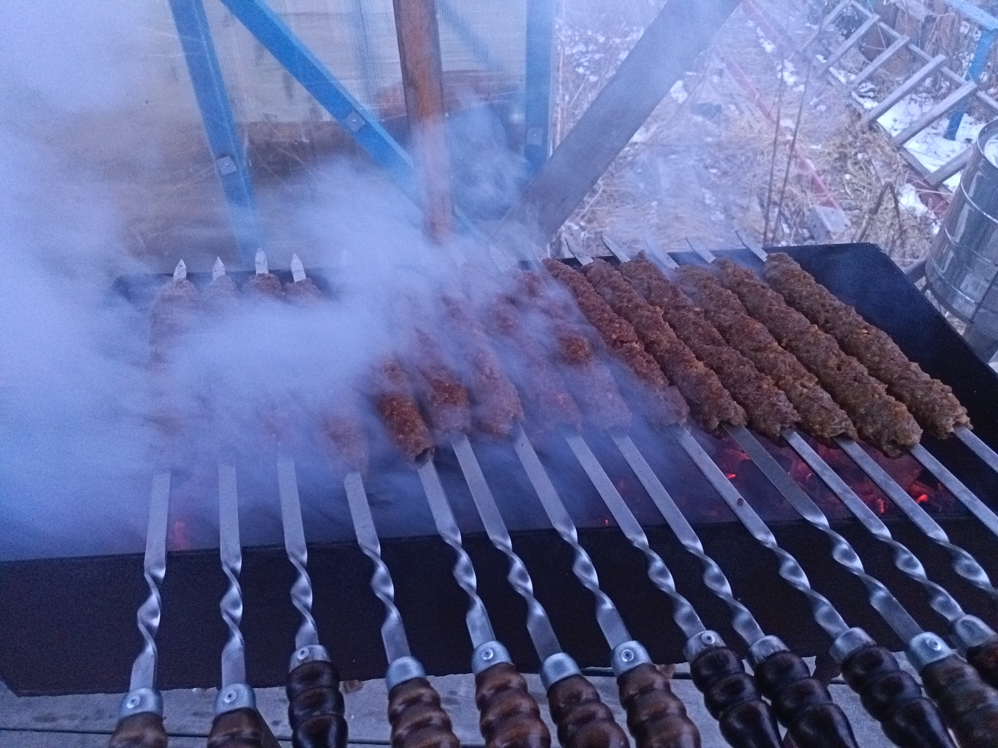 The image depicts a grill with skewers of meat, likely kebab or shashlik, being cooked over hot coals, producing significant smoke. The skewers are arranged in a row over the grill, with their twisted metal shafts and wooden handles visible. The environment appears to be outdoors, possibly near a building or structure with a blue frame. The presence of smoke suggests active cooking.
Given the detail about the city, the scene is potentially taking place in Nizhnevartovsk, Russia.