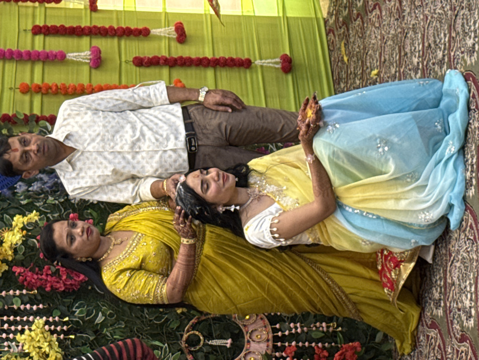 In the city of Malasar, India, a ritualistic scene unfolds indoors. A young adult female, with long dark hair, sits on a small red and gold cushion, facing forward with her eyes downcast. She wears a white blouse with decorative sleeves and a yellow-to-blue ombre saree adorned with white embellishments. Her hands are cupped, holding a yellow granular substance. White paste is visible on her hair and forehead. She has henna applied to her forearms and is accessorized with dangling earrings, a necklace, and a bracelet.

Behind and to her left stands a middle-aged female, dressed in a yellow saree with gold embroidery and matching blouse, accessorized with a heavy gold necklace, earrings, and bangles. Her right hand rests on the seated female's head, indicating an application or blessing. She looks towards the left.

To the right of the seated female, a middle-aged male stands. He wears a light-patterned collared shirt with rolled sleeves, brown trousers, a dark belt, and a silver wristwatch. His right hand is also placed on the seated female's head, near the standing female's hand. He looks slightly to his right.

The background consists of two distinct sections: the left features green foliage adorned with pink and yellow floral garlands and a decorative circular hanging object; the right comprises a bright green fabric backdrop with vertical garlands of red and white flowers. The floor is covered by a patterned rug or carpet with green, red, and brown motifs.