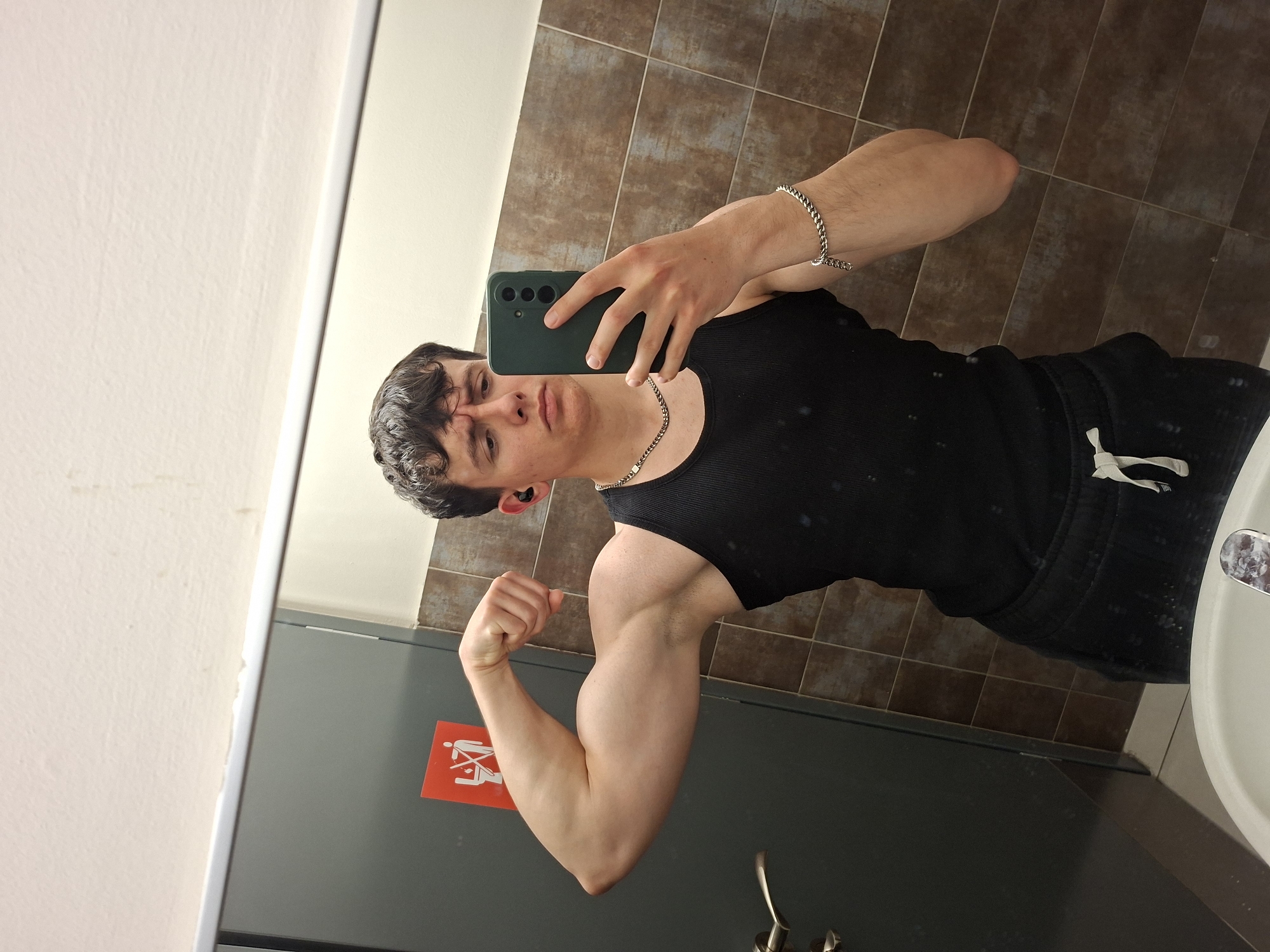 A young male is captured in a mirror selfie, flexing his right bicep in what appears to be a public restroom or changing room in Plovdiv, Bulgaria. He has dark, curly hair and is wearing a black tank top and black sweatpants with a white drawstring. A silver-colored chain necklace and a matching bracelet on his left wrist accessorize his outfit. He holds a dark green smartphone in his left hand to take the photo, and a black earbud is visible in his left ear.
The background shows brown tiled walls, common in public facilities, and a white sink is partially visible on the right. In the reflection, a gray door or cabinet is present on the left side of the frame. A red rectangular sign with a white pictogram is affixed to this gray surface. The pictogram displays two stylized figures, one seemingly pushing another in a wheelchair or stroller, overlaid with a diagonal line, indicating a prohibition or restriction related to the area. The indoor lighting is functional, illuminating the scene. The subject’s expression is focused on posing for the camera, highlighting his muscular arm.