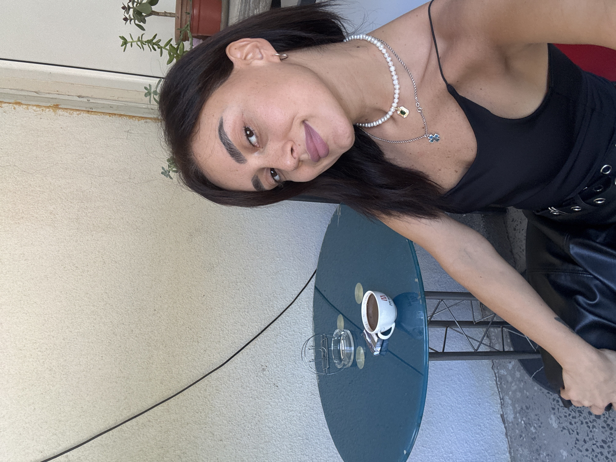 A young woman with dark hair and light skin is sitting at a round, blue table outdoors, in front of a textured, light-colored wall. A cup of dark liquid, likely coffee, sits on the table next to a pack of cigarettes and an ashtray. The woman is wearing a black tank top and several necklaces. Her arm rests on a black leather chair. The overall impression is of a casual, relaxed moment. The location is a street in Tbilisi, Georgia.