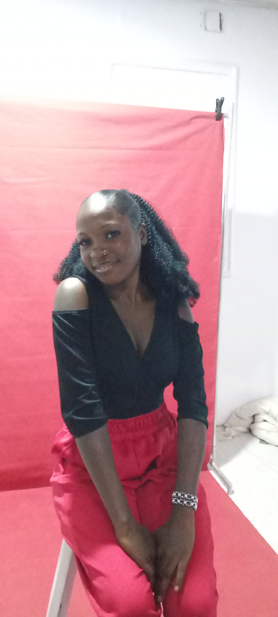 A woman is sitting in front of a red backdrop. She is wearing a black off-the-shoulder top and red pants. She has her hair in a ponytail with curly extensions, and she is wearing a bracelet on her left wrist. The background appears to be a makeshift studio setup. The location is Gura Top, Nigeria. The scene is a portrait-style photograph.