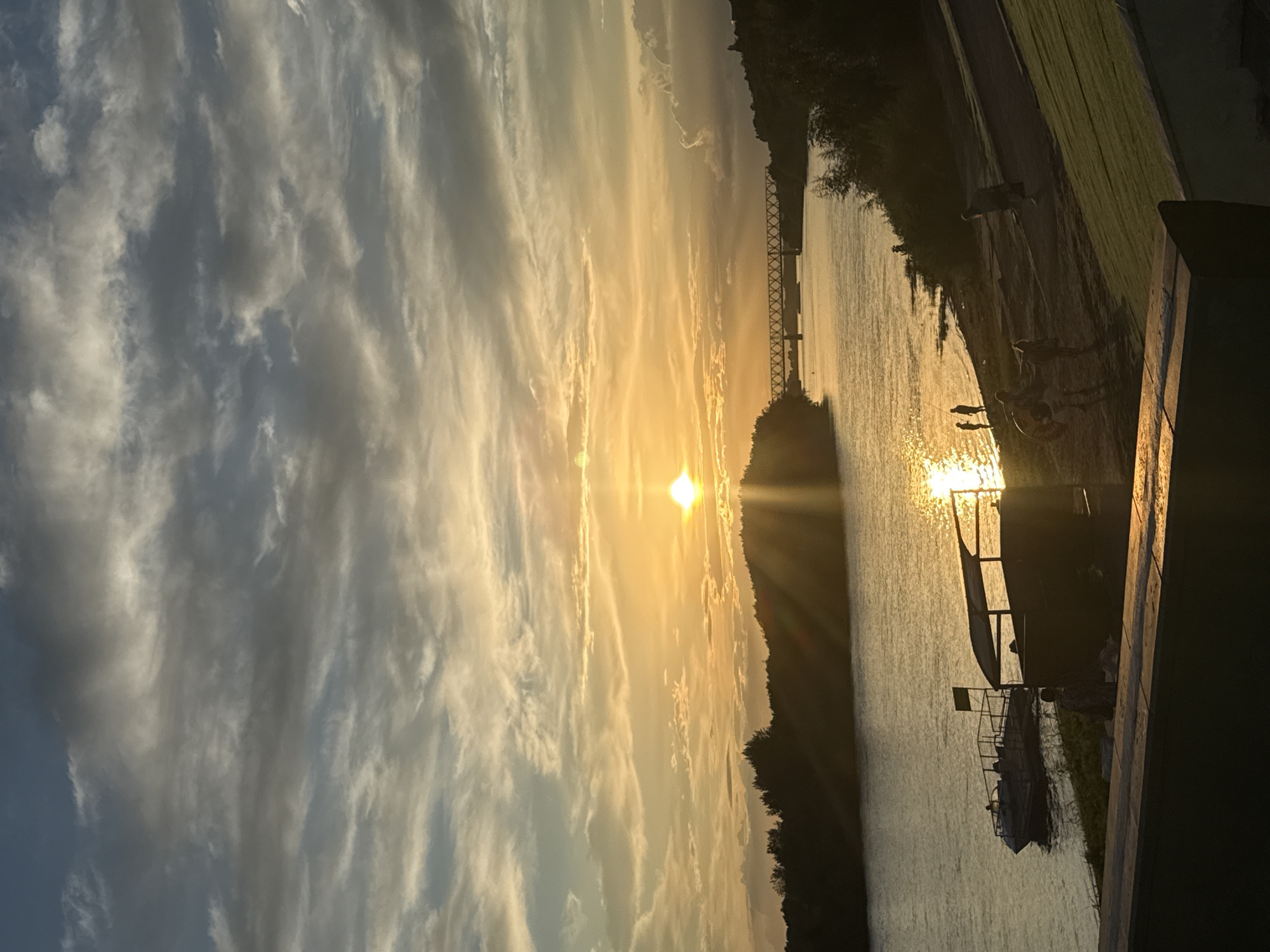 The image depicts a serene sunset over a body of water, likely a river, in Pavlodar, Kazakhstan. The sky is filled with dramatic, streaky clouds painted in hues of orange, yellow, and gray, reflecting the sun's warm glow. The sun itself is a bright, small orb just above the horizon, casting long rays of light across the water.
On the right side of the image, a tree-lined shore is visible, silhouetted against the setting sun. A walkway or embankment runs along the water's edge, with a few people standing or sitting, enjoying the view. Further along the shore, a dark structure, possibly a pier or a boat dock, extends into the water, with two boats moored alongside it. One boat is more prominent, with a canopy.
The overall mood of the image is peaceful and contemplative, capturing the beauty of a natural sunset. The time of day is clearly evening, as indicated by the low position of the sun and the warm, golden light. The weather appears calm, with the water being relatively still. There is no visible text in the image.