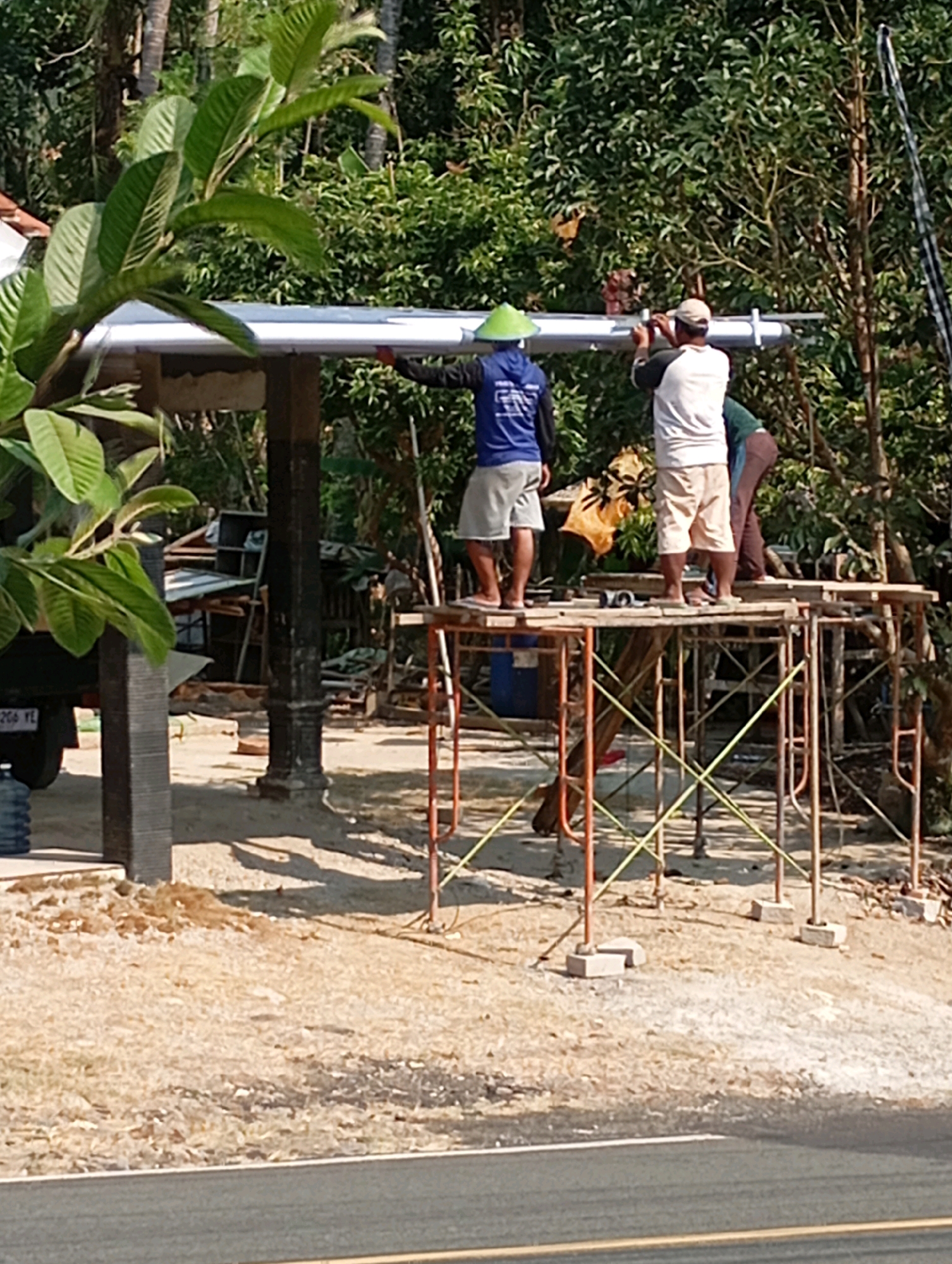 The image depicts a construction scene in Ketapang, Indonesia, where three men are working on installing a roof structure. They are positioned on a scaffolding system erected on a sandy, outdoor area, with a road visible in the foreground. The environment is rural or semi-rural, characterized by abundant trees and foliage in the background, suggesting a tropical or subtropical climate.
One man, wearing a blue shirt and a conical green hat, is seen on the left side of the scaffolding, assisting with the roof installation. To his right, another man in a white and brown shirt and a cap is actively engaged in securing the roof. A third individual, partially visible behind the man in the white shirt, appears to be providing support.
The scaffolding is made of metal poles and planks, with yellow and orange accents. Beneath the scaffolding, a large blue plastic barrel is present, along with various construction materials and debris, hinting at ongoing work. A truck is parked to the left of the scene, partially obscured by foliage. The overall impression is one of manual labor and construction in a natural, sunlit setting.