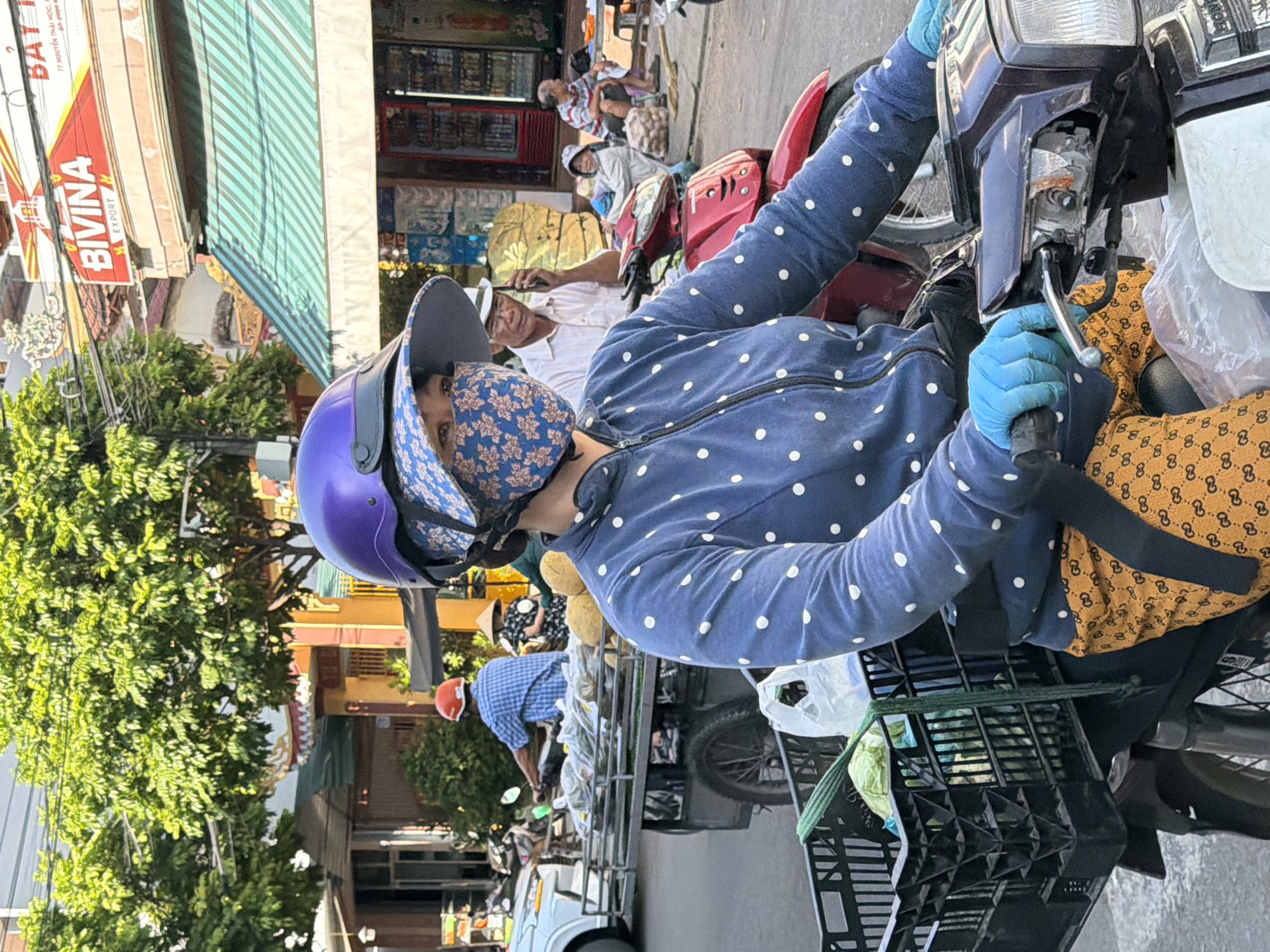 A woman wearing a blue polka dot jacket, blue gloves, a floral face mask, and a purple helmet rides a motorcycle in Nha Trang, Vietnam. The motorcycle has a black basket attached to the front, filled with produce. She is wearing yellow patterned pants. In the background, a man in a blue checkered shirt rides a motorcycle pulling a cart filled with produce. Other people and motorcycles are visible on the street. Buildings line the street, some with awnings and signs. The environment appears to be a busy urban street.