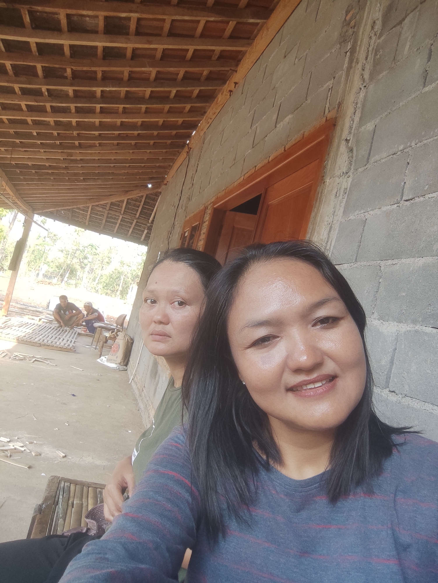 The image is a selfie taken outdoors in Mojopuro, Indonesia, showing two women in the foreground. The woman on the right is closer to the camera and is smiling. She has dark hair and is wearing a blue and red striped t-shirt. The woman on the left is slightly further back and looking off to the side. She has dark hair and is wearing a green t-shirt. Behind them, there is a building with a concrete block wall and a wooden roof structure. To the left of the building, two men are visible in the background, one sitting and the other kneeling, working on what appears to be bamboo structures. The ground is dirt or gravel. The environment suggests a rural or semi-rural setting, possibly a construction site or a workshop.