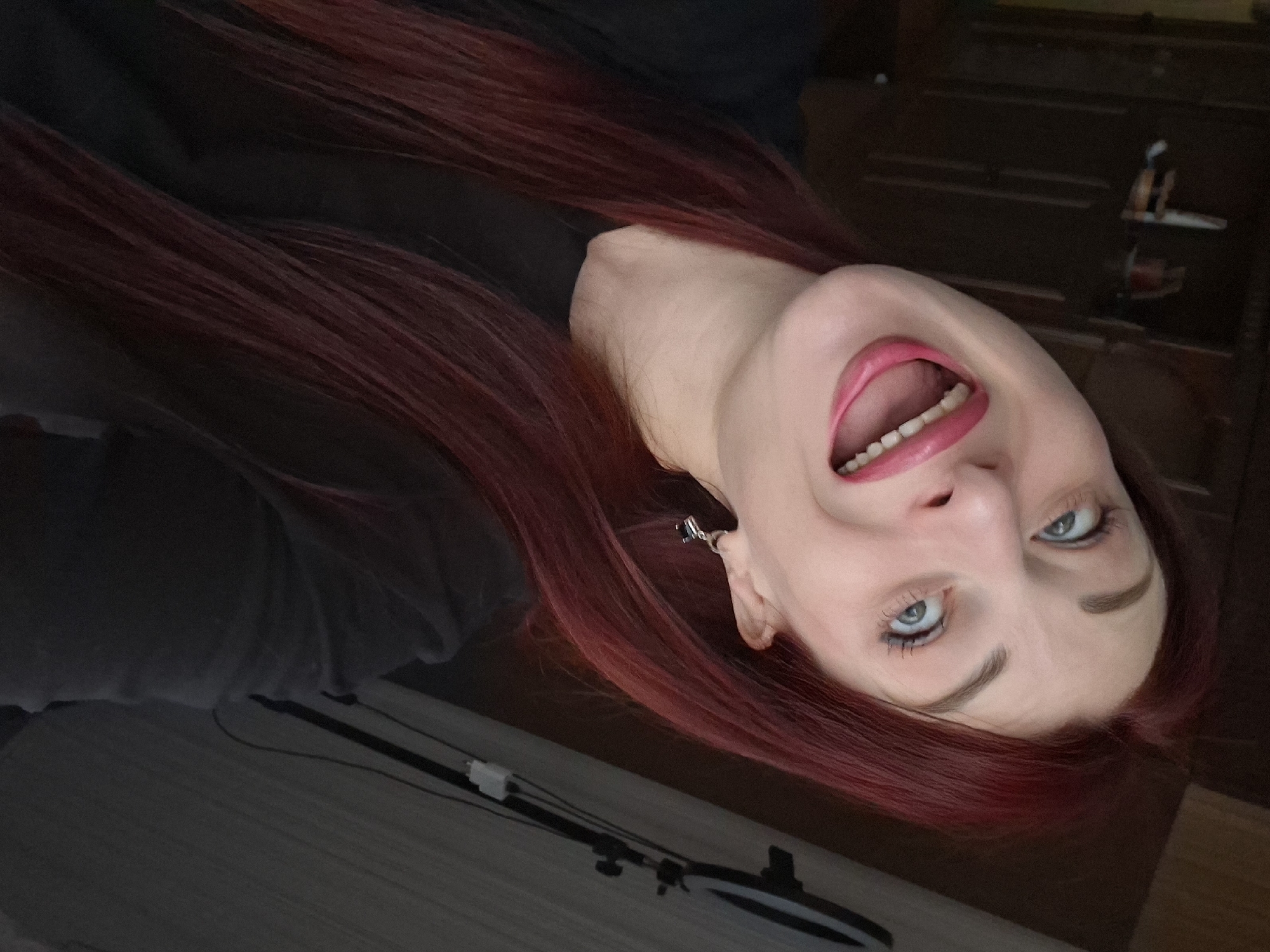 The image is a close-up selfie of a woman with red hair, fair skin, and light blue eyes. She has bright pink lipstick on and her mouth is open. She is wearing a dark colored shirt. There is an earring visible in her left ear. The background appears to be a room with dark wood furniture. There is a ring light visible in the bottom left of the frame. The location in the city is Unknown, Unknown.