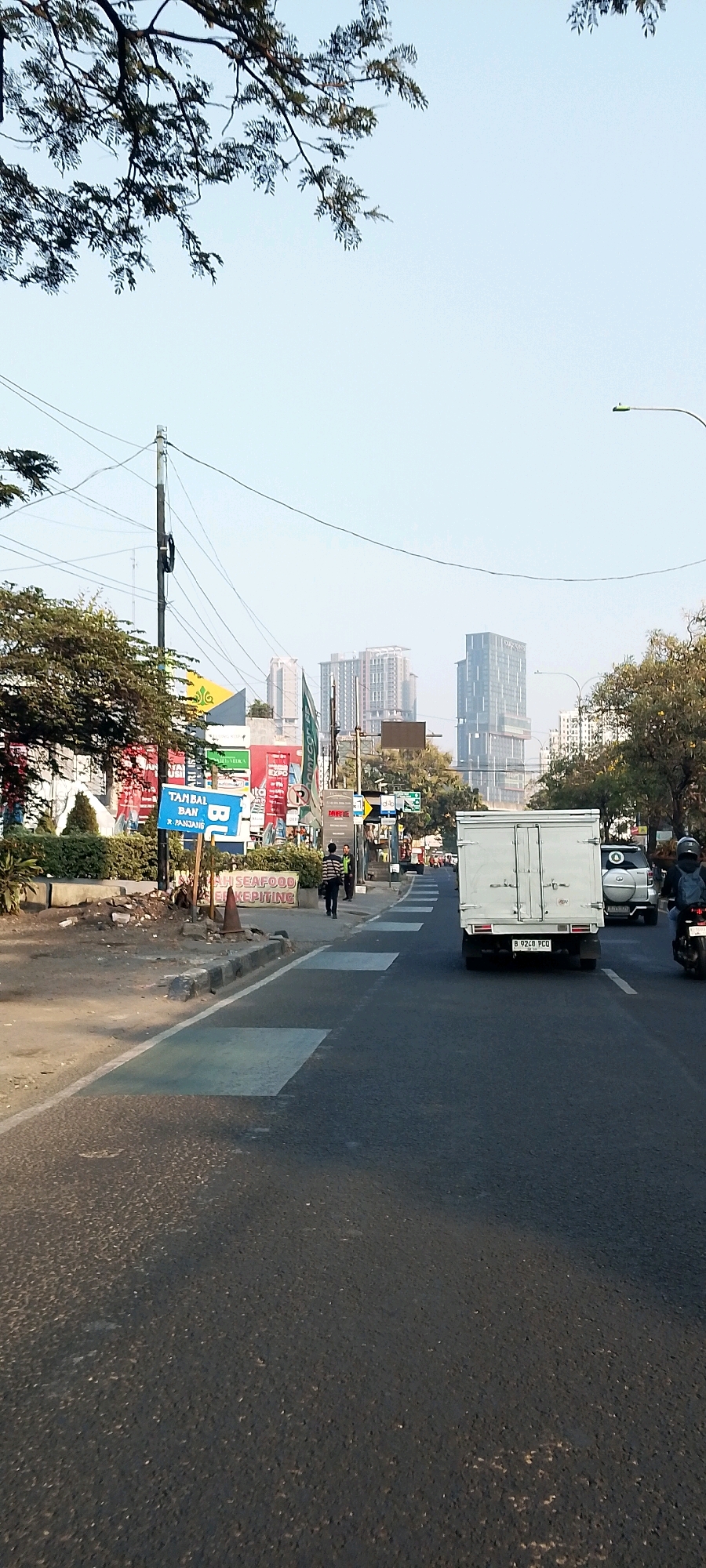 The image depicts a street scene in Bekasi, Indonesia, under a clear sky. The foreground shows an asphalt road with painted crosswalks. A white box truck with the license plate "B 9248 PCO" is traveling away from the viewer in the right lane. To the right of the truck, a silver SUV and a motorcycle with a rider wearing a backpack are also moving away. On the left side of the road, there are various advertisements and signage, including a blue sign that reads "TAMBAL BAN" and "R. PANJANG". A pedestrian in a striped shirt is walking on the sidewalk. In the background, several tall modern buildings dominate the skyline, indicating an urban environment. Trees line the sides of the road, and overhead power lines are visible.