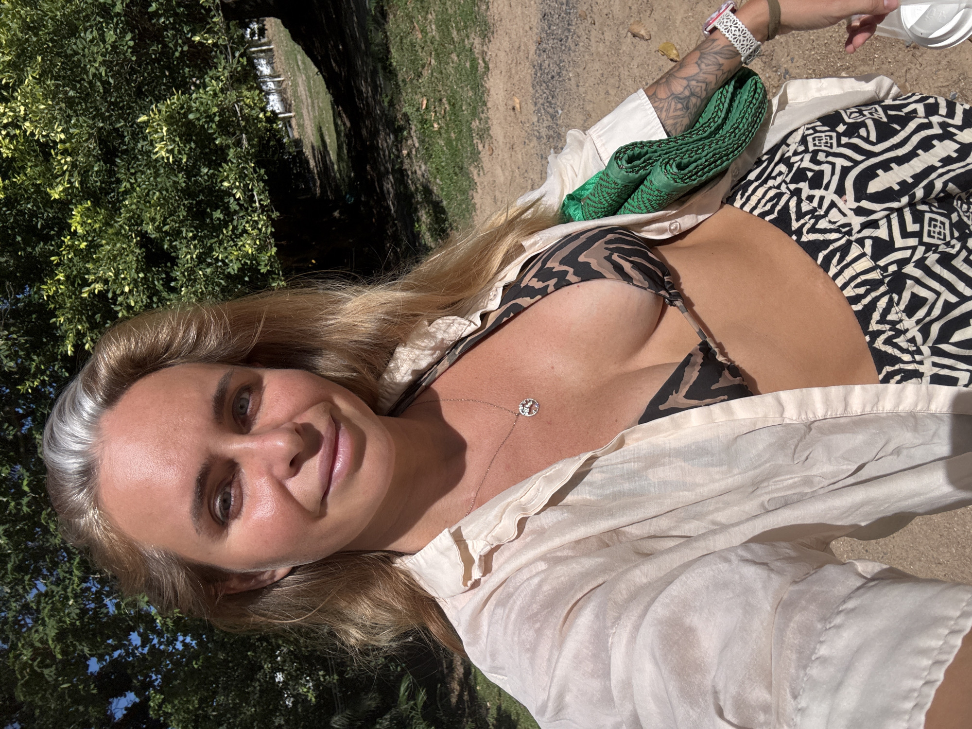 A woman stands outdoors, in what appears to be a park or natural area, possibly in Baan Chaweng Noi, Thailand. She is facing the camera and smiling, with her left arm extended to take the photo. She is wearing a tan and black patterned bikini top, a loose-fitting, light-colored unbuttoned shirt, and patterned pants. A green rolled-up item, possibly a mat or blanket, is held under her right arm. Her hair is blonde and wavy, with some grey streaks visible. The background features lush green trees and dappled sunlight.